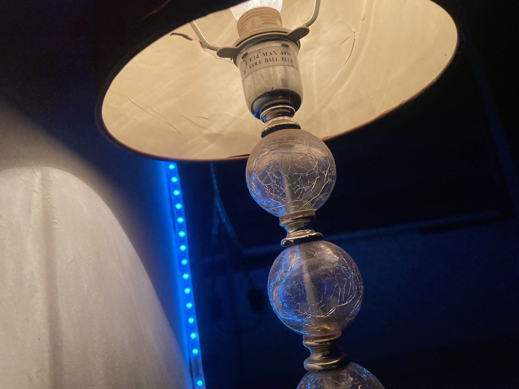 The image depicts a lamp with a tan shade and a stacked, cracked-glass ball base. The lamp is lit, revealing the bulb fitting labeled "E14 MAX 40W GOLF BALL BULB". A strip of blue LED lights is visible in the background. A portion of a white surface is also visible on the left. The location is not discernible from the image.