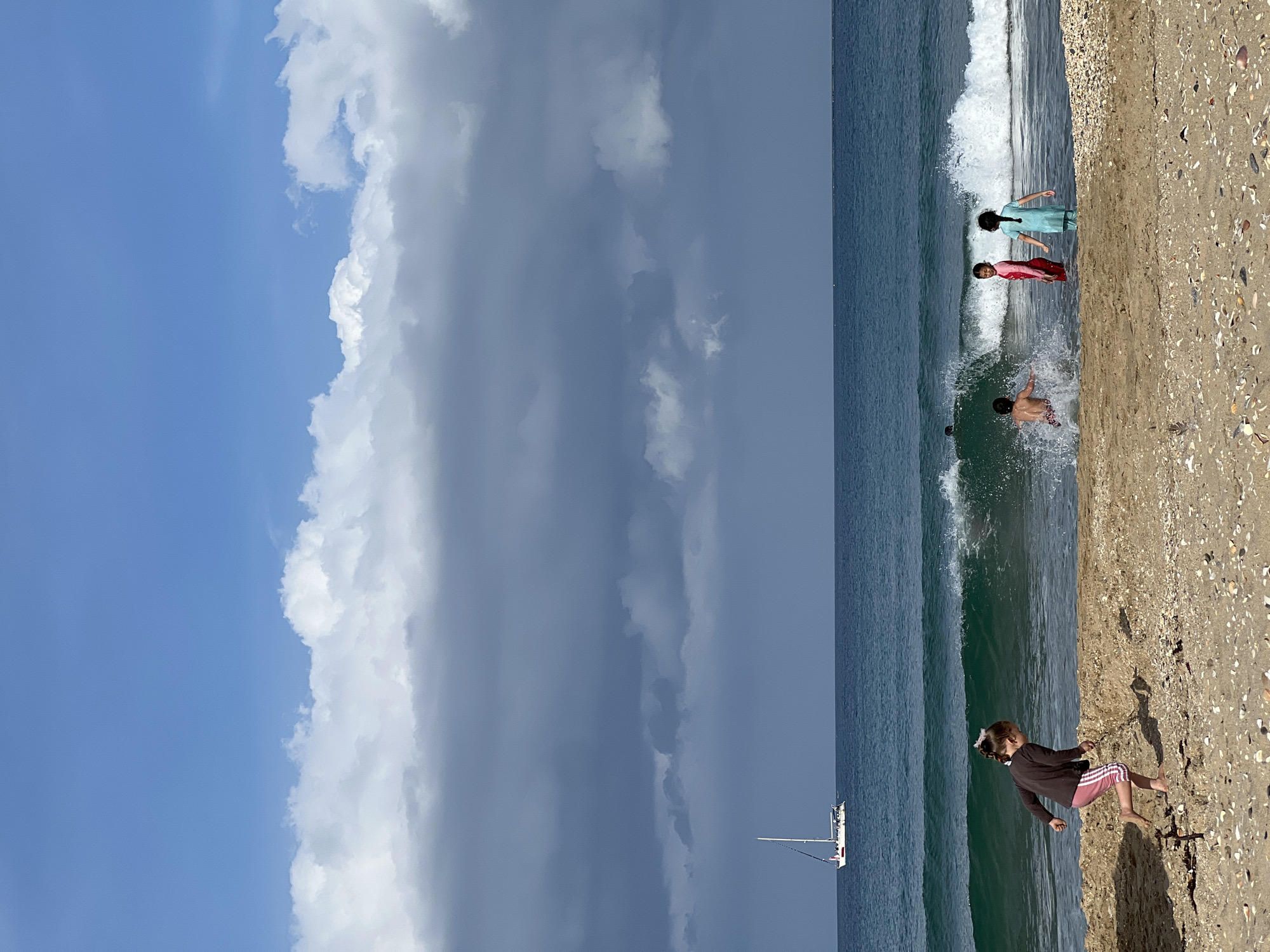 The image depicts a beach scene in M'Diq, Morocco. The sky is partially cloudy, with a mix of blue sky and dark rain clouds. The ocean meets the sandy beach, where several people are present. Three individuals are partially submerged in the water, seemingly playing in the waves. On the beach, a young child is standing near the water's edge. A sailboat is visible on the water in the distance.