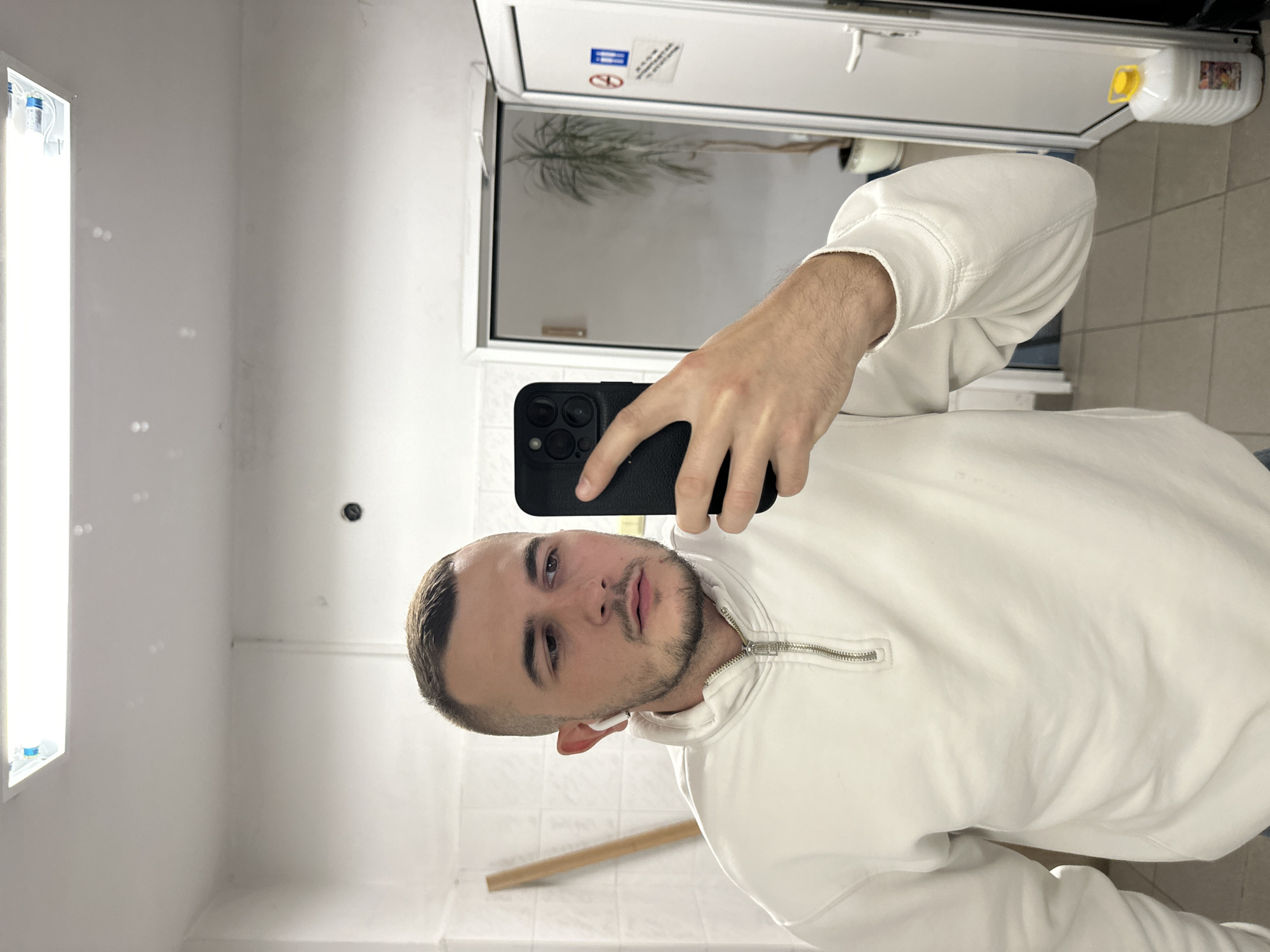 The media file displays a selfie taken by a young man indoors, with the image rotated 90 degrees counter-clockwise. The man, likely in his 20s, has short dark hair, a trimmed beard, and a light mustache. He wears a white or off-white hooded sweatshirt with a silver zippered collar and has a white earbud in his right ear. His left hand holds a black smartphone, featuring multiple camera lenses, which he uses to take the picture. He looks directly at the camera with a neutral expression.
The setting appears to be a brightly lit, utilitarian interior, possibly a hallway or utility room in Plovdiv, Bulgaria. The walls and ceiling are plain white, with a long fluorescent light fixture mounted vertically on the left side of the frame, showing small blue bottles or objects inside. A dark circular object, possibly a sensor or camera, is visible on the upper left wall. The lower wall and floor are covered with light-colored tiles, and a light brown wooden stick leans against the wall in the bottom right.
Behind the man, a white door with a reflective window section reveals a blurred reflection of green foliage, possibly a potted plant. On the upper part of the door, there are several white stickers, one with a blue background and another featuring a crossed-out symbol, possibly indicating a prohibition like "no smoking." A white plastic bottle with a yellow cap and an illegible colorful label rests on a ledge above the door.