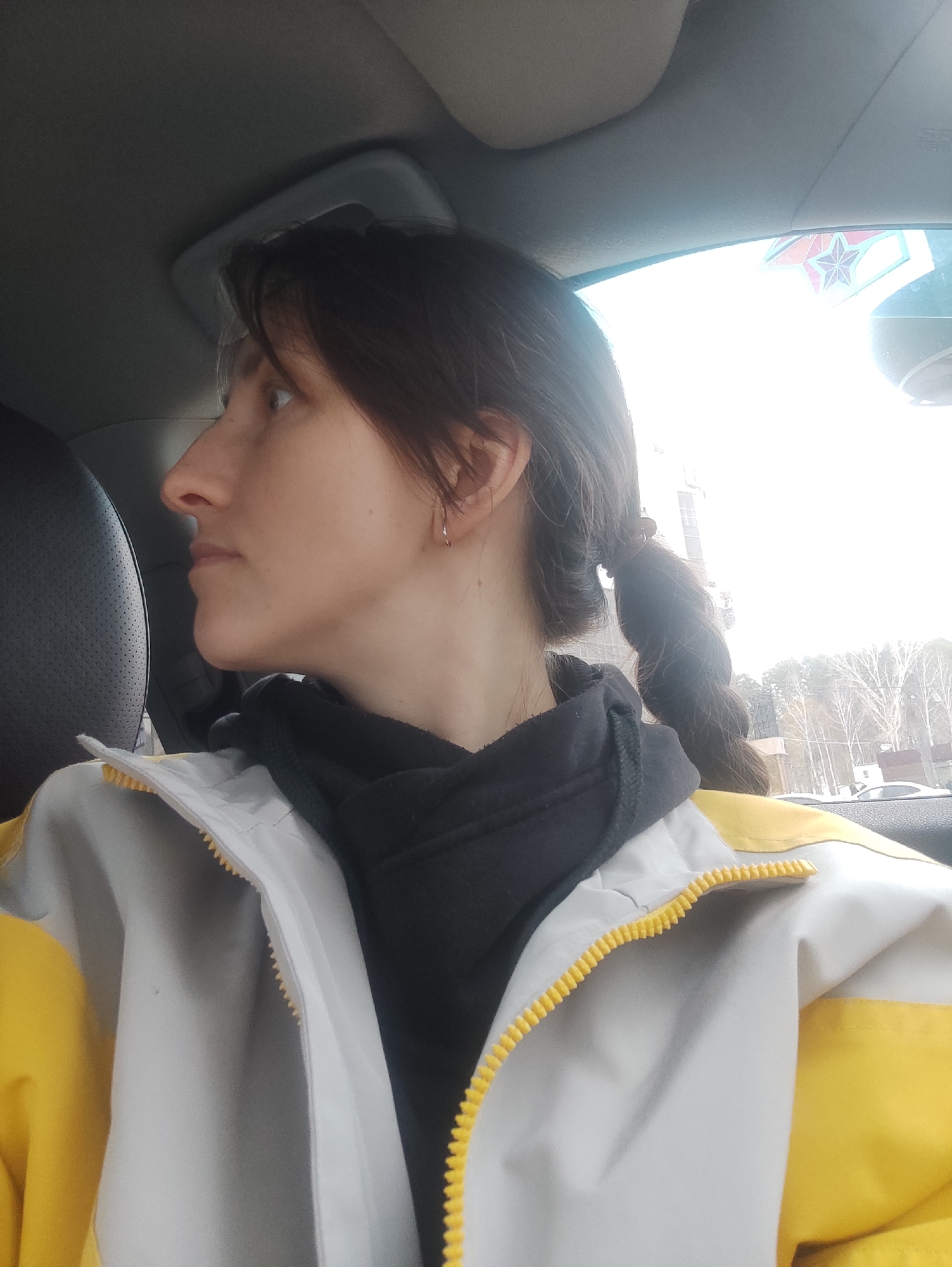 A female subject is seated in what appears to be the front passenger seat of a vehicle in Chaikovsky, Russia. She is depicted in profile, looking towards the right side of the frame. The subject has dark hair, styled into a low ponytail or braid visible over her right shoulder, and a fair complexion. She wears a small, silver-colored hoop earring in her left ear. Her attire consists of a black hooded garment worn under a two-tone jacket, which is light grey on the main body and yellow on the shoulders and arms, featuring a yellow zipper closure.
The vehicle's interior is visible, including a grey headliner and a black textured seat to the left. Through the vehicle's window, an outdoor environment is discernible under bright, diffused daylight. Bare deciduous trees, characteristic of a colder climate or season, are visible. In the background, there are buildings, including a taller, multi-story structure, and other vehicles. A prominent red star emblem, likely part of a monument or architectural feature, is partially visible through the upper right portion of the window. The subject's action involves observing the external environment.