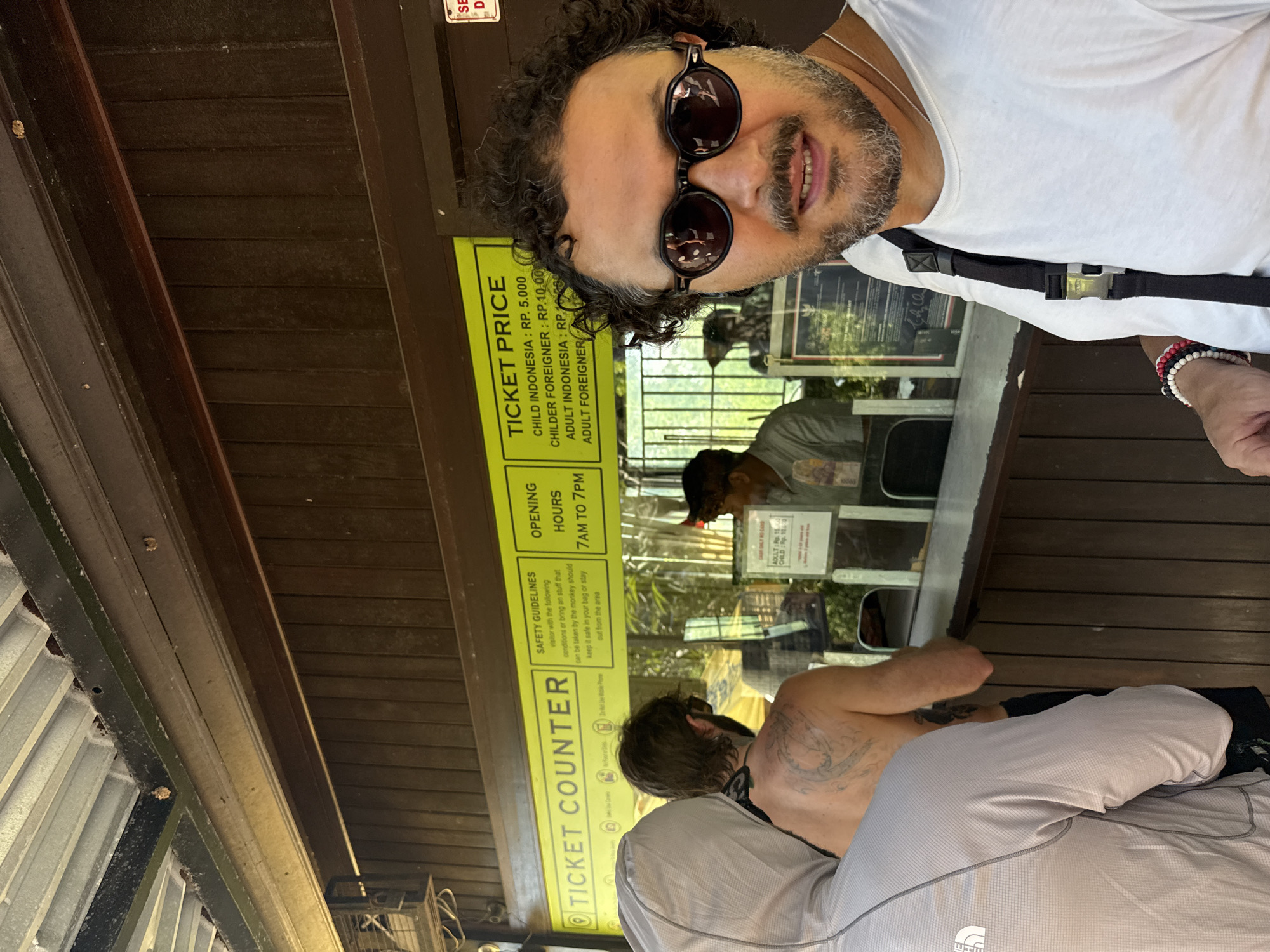 A man with curly hair and sunglasses poses in front of a ticket counter. The ticket counter lists prices for child and adult tickets for Indonesian citizens and foreigners, as well as the operating hours of 7 AM to 7 PM. It also contains safety guidelines regarding monkeys. A person is at the counter, and another person is seated. Based on the ticket price, this is likely near a tourist attraction. A partially legible sticker on the ticket window reads, "SE D." It is difficult to determine the exact location within Kuta Selatan.