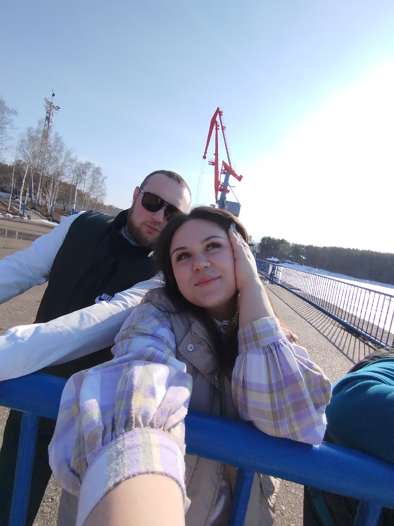On a sunny day in Chaikovsky, Russia, a man and a woman are standing on a blue-railed bridge, posing for a selfie. The man, wearing a white long-sleeved shirt under a black vest and sunglasses, leans in from the left side of the frame. He has a dark beard. The woman, positioned closer to the camera, is wearing a light-colored jacket with a plaid-patterned sleeve. She is resting her hand on her face.
Behind them, a red crane structure rises to the left, while the bridge spans a waterway bordered by a snowy bank and a line of trees. A light-colored tower stands near a stand of bare trees to the left. The bright sunlight fills the scene.