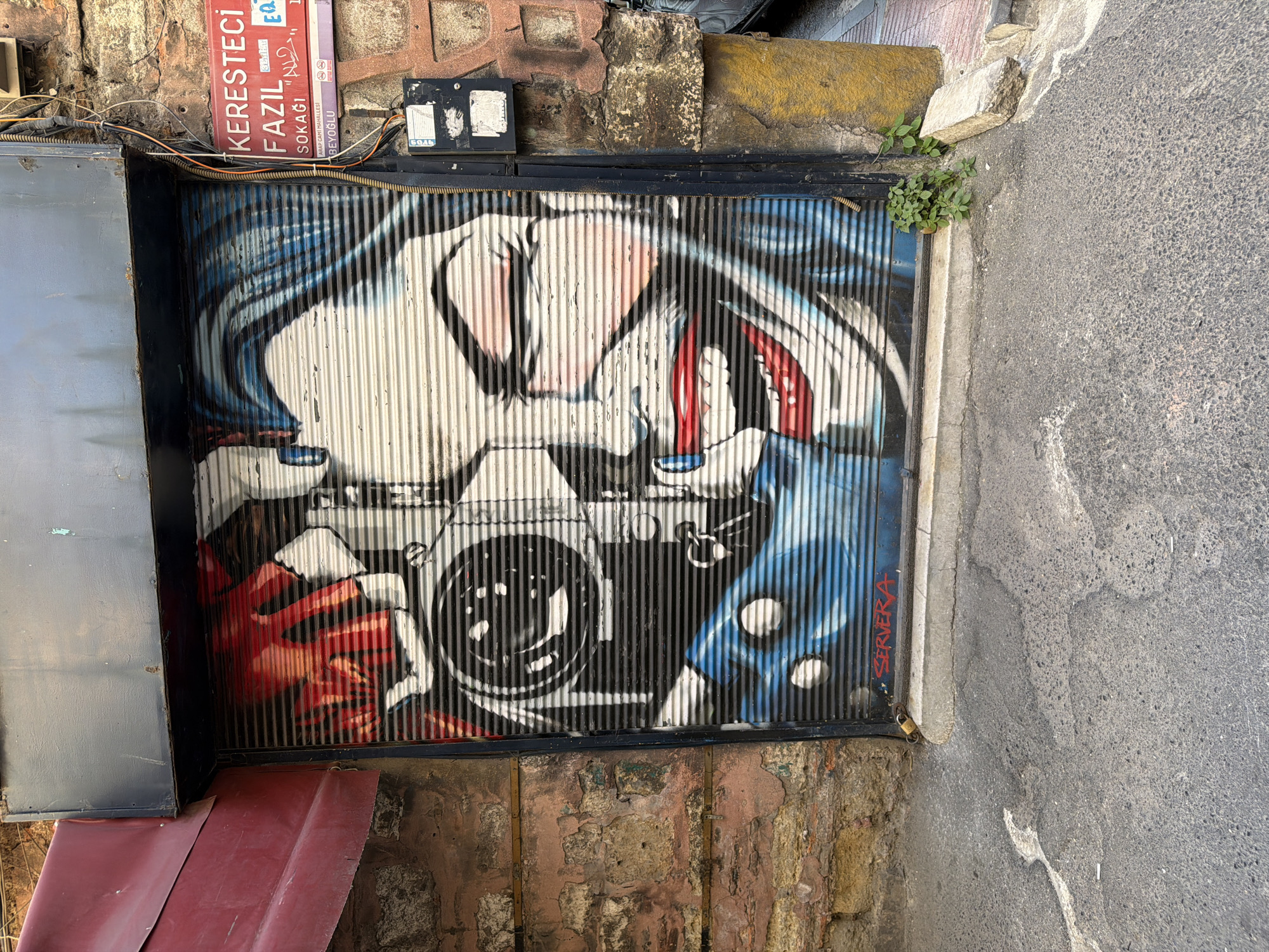 This is a photograph of graffiti art on a metal security shutter in the city of Beyoğlu, Turkey. The artwork depicts a close-up of a face, possibly inspired by the Joker character from Batman, with a camera lens integrated into the face. The face is rendered in bold colors, with prominent red lips, white skin, and blue and black accents for the hair and shadows. The security shutter itself is made of horizontal metal slats, which create a corrugated effect that is visible through the graffiti. To the right of the shutter, there is a brick wall with a street sign indicating "KERESTECI FAZIL SOKAĞI," which translates to "Keresteci Fazıl Street" in Beyoğlu. Electrical conduits and a small electrical box are also attached to the wall. To the left of the shutter, a red awning extends outwards, casting a shadow. The ground in front of the shutter is asphalt, with some cracks and patches. A small plant is growing near the base of the shutter, and a broken piece of concrete lies on the ground next to it. A padlock is visible on the left side of the shutter, securing it. The overall scene suggests an urban environment, likely a shop entrance.