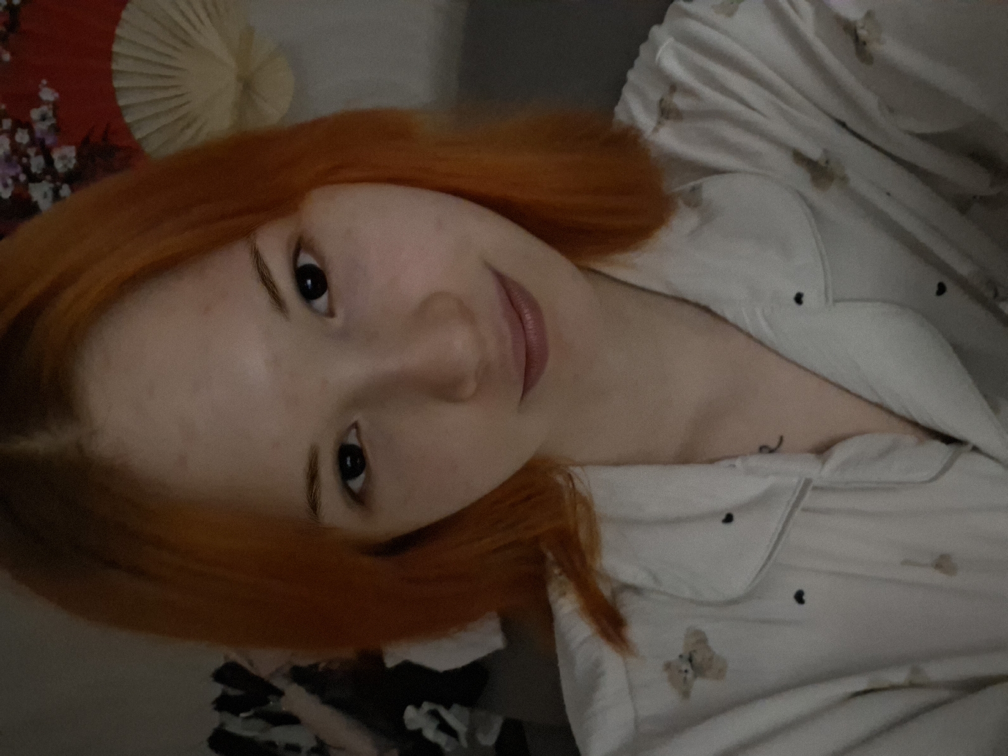 A young woman with bright, reddish-orange hair and fair skin is captured in a close-up, horizontal portrait, seemingly lying down indoors in Chaikovsky, Russia. She is looking directly at the camera with a soft, gentle smile, her dark eyes prominent.
She is wearing light-colored pajamas, likely white or cream, featuring small, scattered dark heart patterns and light brown teddy bear motifs. A faint, dark, indistinct marking, possibly a small tattoo, is visible on her upper chest area, near the collarbone, partially covered by her pajama top.
In the background, positioned above her head, are two decorative fans: one is predominantly red with delicate white floral or cherry blossom patterns, and the other is a lighter, pleated fan, possibly beige or cream. The lighting is soft and ambient, suggesting an indoor setting during evening or night hours, contributing to a relaxed and calm atmosphere. There is no visible text in the image.