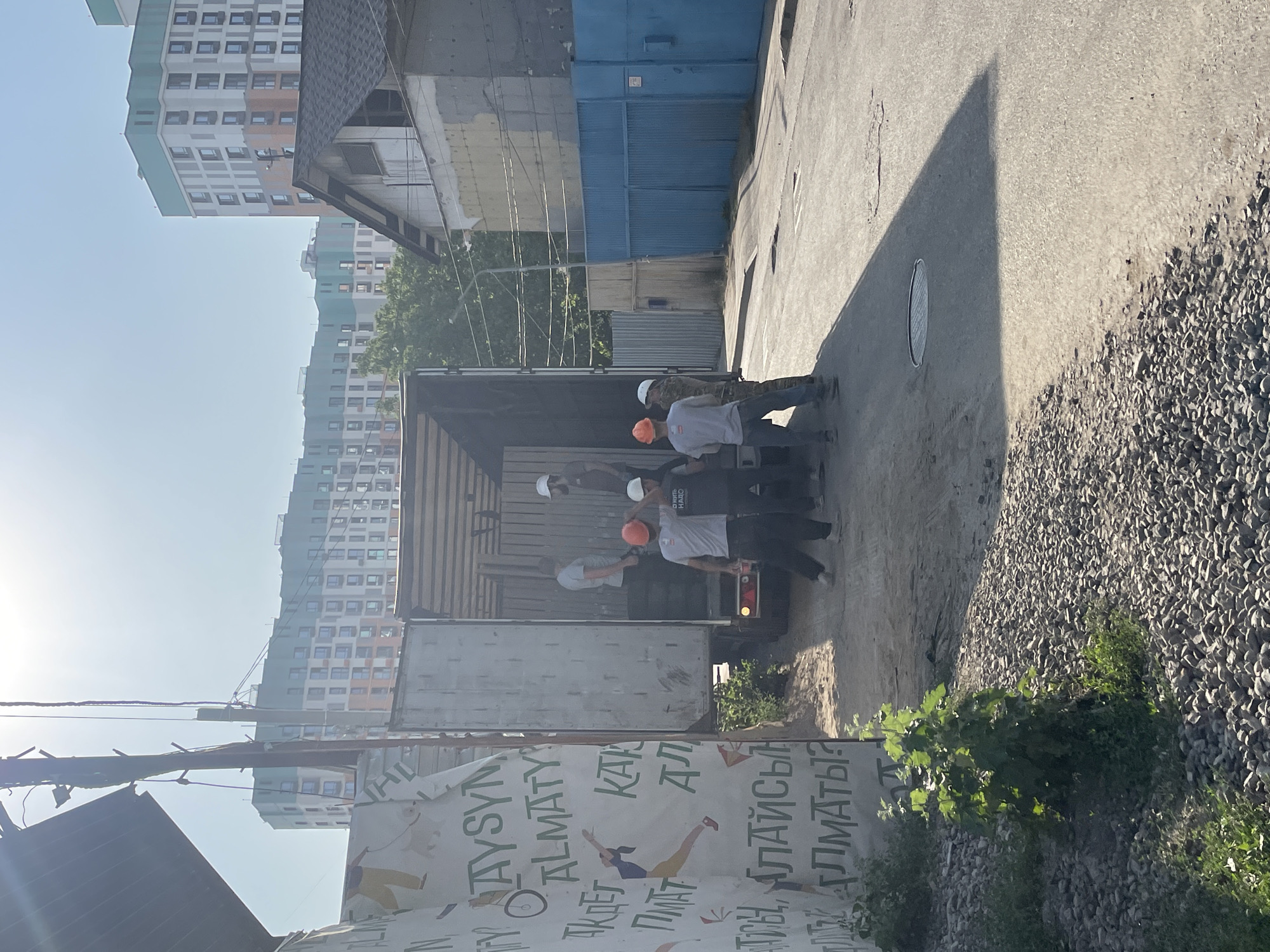 The image depicts a scene in the city of Almaty, Kazakhstan, on a bright, sunny day. In the foreground, a truck with its rear doors open is being loaded or unloaded by a group of construction workers. Five men, all wearing hard hats, are visible. Three are standing near the truck, and two are inside it, seemingly engaged in the task. The truck is parked on a paved road, with a section of gravel and a small bush to the left. In the background, several modern, multi-story buildings with light-colored facades and green accents rise against a clear blue sky. A blue metal fence runs along the right side of the road, adjacent to a building. A large banner with text and graphics is partially visible on the left, leaning against a structure. The overall atmosphere suggests ongoing construction or a logistical operation in a residential or mixed-use urban area.
