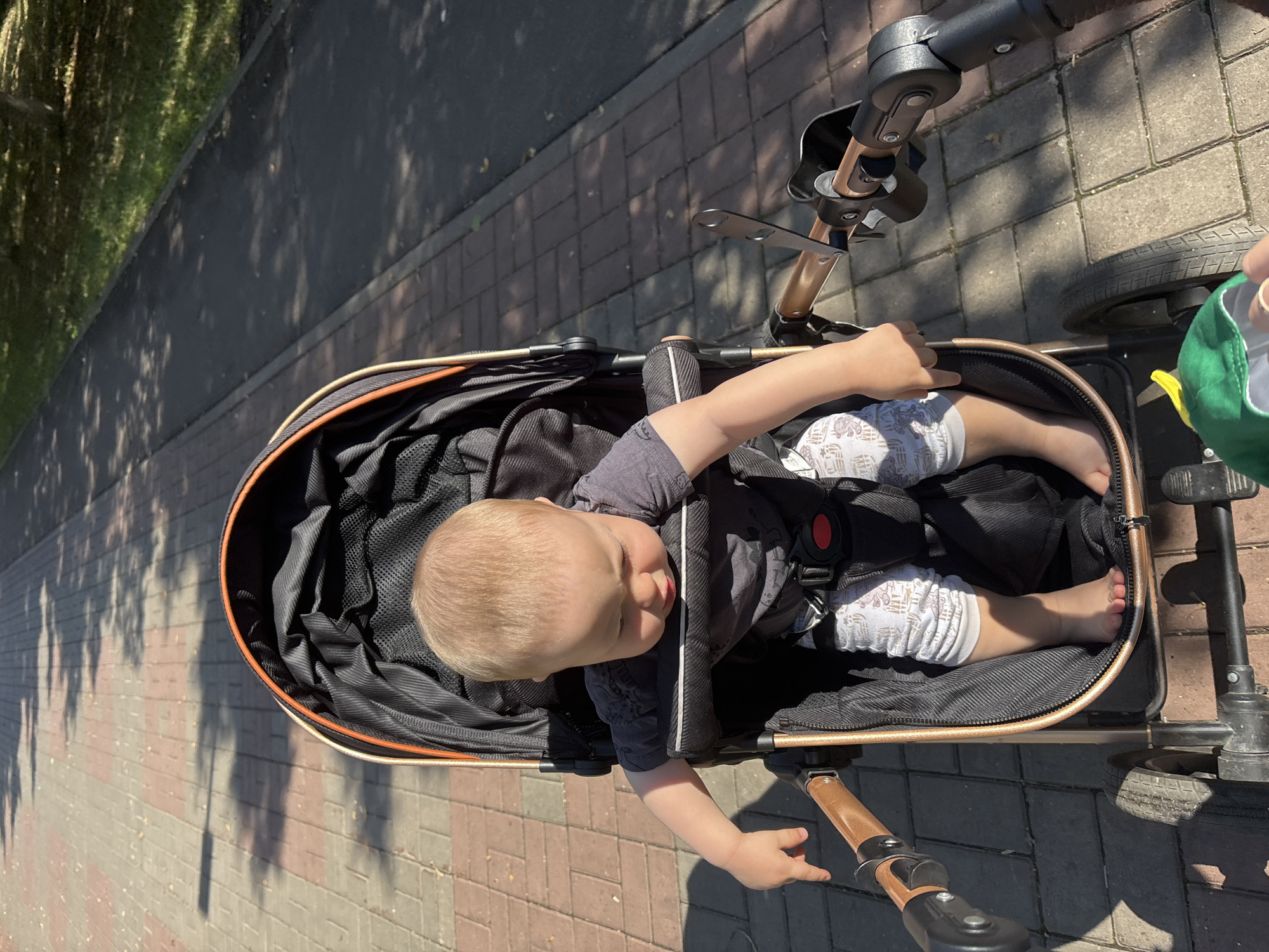 A young, fair-haired child is seated in a stroller on a paved path. The child is wearing a dark gray t-shirt and patterned leggings. The stroller is black with gold accents and has a red buckle. The child is looking down and to the side, with their left hand resting on the edge of the stroller. Sunlight casts shadows on the path, which is made of reddish and gray rectangular pavers. A patch of green grass and trees is visible to the right of the path. The scene appears to be in an outdoor public area, possibly a park or a residential street. This image was captured in Naberezhnye Chelny, Russia.