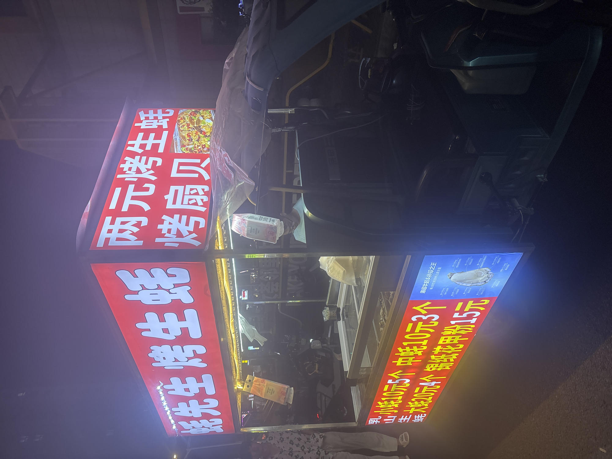 A night-time street food vendor is parked on a paved surface, likely a sidewalk or street, in Suzhou Industrial Park, China. The vendor is a three-wheeled vehicle with a prominent, illuminated red sign displaying Chinese characters for "Mr. Crab Grilled Oysters" and "Two Yuan Grilled Oysters" along with an image of a dish. The sign also lists prices for different items such as "Small Oysters 10 Yuan for 5," "Medium Oysters 10 Yuan for 3," and "Large Oysters 20 Yuan for 4." Another section of the sign, on the right, shows an image of an oyster with nutritional information in Chinese. A person in a patterned shirt and light-colored pants stands to the left of the vendor. The vendor's stall has a glass display case that appears to contain raw oysters and other seafood. A tarp is draped over the top of the vehicle. The surrounding environment is dark, with some ambient lighting from streetlights or other signs visible in the background.