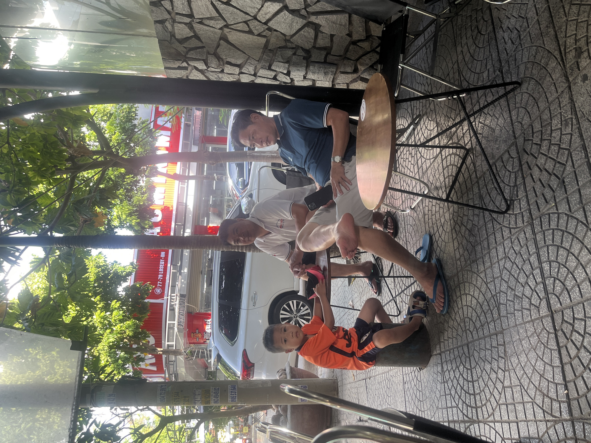 This detailed image depicts an outdoor street scene in Đà Nẵng, Vietnam, specifically identifiable by the "77-79 LOSEBY-BN" sign on a red building in the background. The foreground features a paved sidewalk with grey patterned tiles, where three Asian individuals are seated under the partial shade of trees.
On the left, a young boy, appearing to be 5-8 years old, is dressed in an orange and black short-sleeved shirt and matching shorts. He sits on a low, dark cylindrical stool, holding a pink object in his hands, and looks directly towards the viewer with a neutral expression. He wears blue and white sandals.
To the boy's right, an adult man, likely middle-aged, wears a white polo shirt with blue trim and light-colored shorts. He is seated at a small, round wooden-topped table with a black metal base, casually crossing his left leg over his right, and appears to be looking towards the left. He wears black flip-flops.
Further to the right, another adult man, also middle-aged, is seated in a black foldable chair against a textured wall composed of irregular grey and brown stones. He is dressed in a dark blue polo shirt with white trim and light-colored pants. He holds a dark-colored smartphone in his left hand, which rests on his lap, and wears a silver watch on his left wrist. His gaze is directed towards the left side of the frame, past the first man, with a calm expression.
In the background, a white Mitsubishi Outlander SUV is parked on the street. Behind it, a prominent red building with white lettering displays "77-79 LOSEBY-BN" and a "San Miguel" beer advertisement in a red barrel shape. Several trees with vibrant green foliage frame the upper part of the image, providing dappled sunlight. Bright sunlight from the upper left casts distinct shadows across the scene. On the far left, a utility pole is visible, adorned with various faded notices and phone numbers, including "0934 72 5972" and "09634949 82". The scene portrays a casual, relaxed urban environment, likely a street-side cafe or gathering spot.