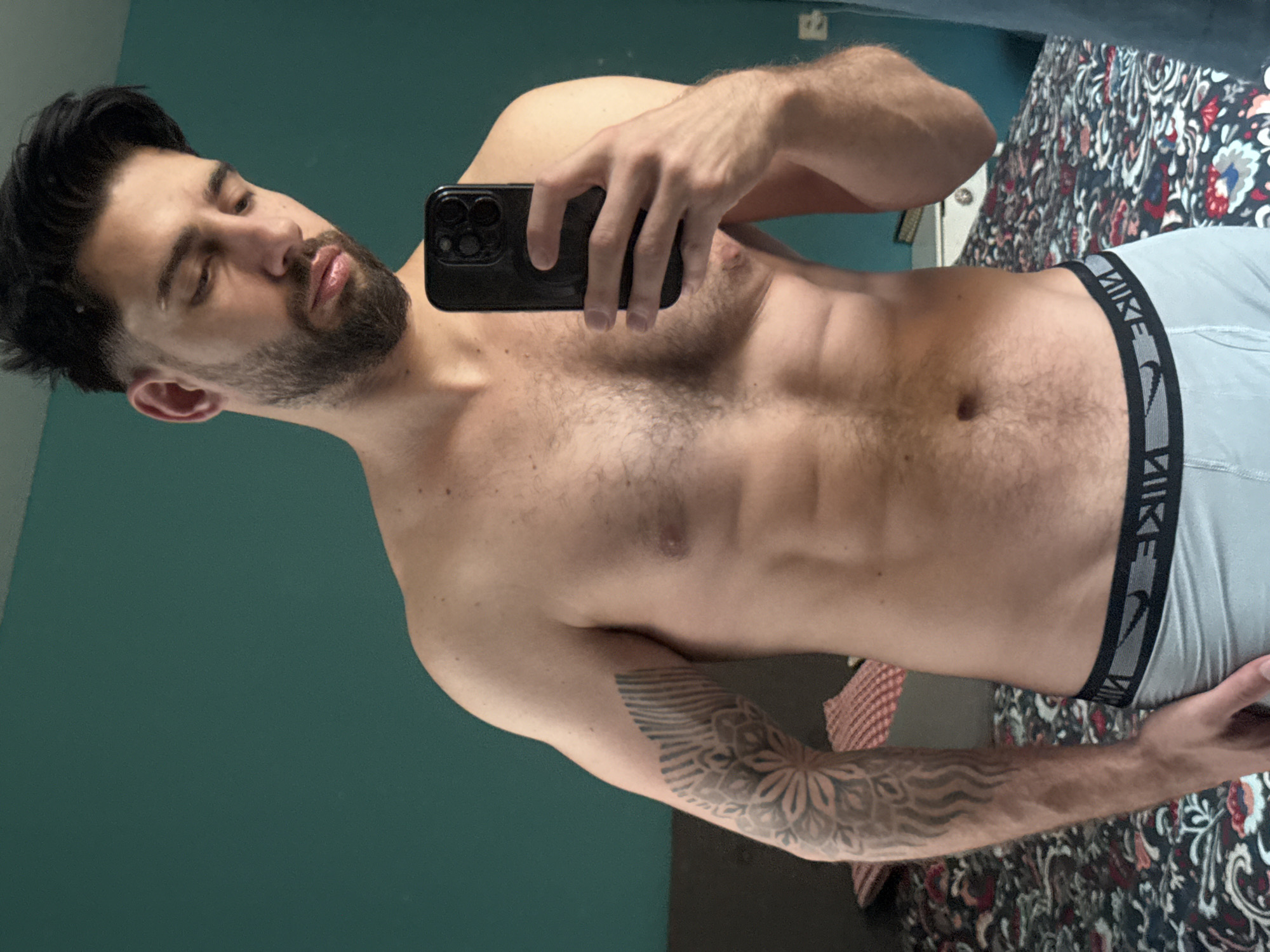 The image features a man taking a mirror selfie indoors in Sofia, Bulgaria. He is shown from the chest up, with his torso slightly angled. The man has dark hair and a well-groomed beard, appearing to be in his 20s or 30s. His eyes are partially closed or looking downwards, and his lips are slightly parted. He possesses a muscular physique with visible chest hair and defined abdominal muscles.
He is holding a black smartphone, identifiable by its prominent triple camera array, in his right hand, which partially covers his upper torso. An elaborate sleeve tattoo, featuring intricate geometric and floral patterns, is visible on his left forearm.
The man is wearing light gray boxer briefs with a black elastic waistband that prominently displays the word "NIKE" in white capital letters, along with the brand's iconic swoosh logo.
The background includes a solid teal or dark green wall on one side. On the other side, a patterned fabric, possibly a bedspread or upholstery, is visible, featuring a dark base with white, red, and blue floral or paisley-like designs. The lighting suggests an interior setting. The image is rotated counter-clockwise.
