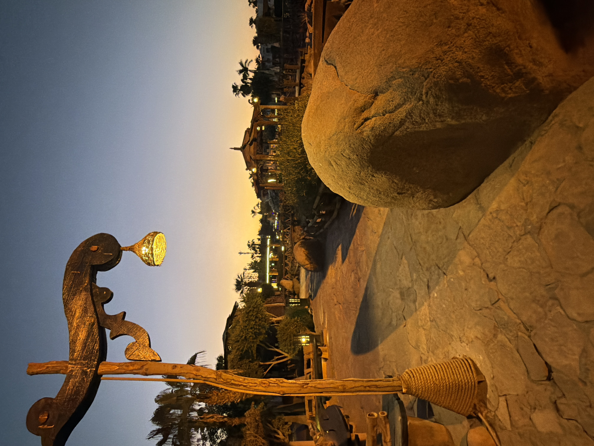 The image depicts an outdoor pathway in Sharm El Sheikh, Egypt, at twilight. In the foreground, a large, rounded rock is illuminated by warm light, casting a shadow on the stone-paved path. To the left, a wooden lamppost with an ornate, curved arm holds a woven, conical lampshade that emits a soft, yellow glow. The base of the lamppost is wrapped in coiled rope. Further along the path, a wooden table and benches are visible, suggesting a seating area.
In the midground, the pathway leads towards a building with a tiered, wooden roof structure, possibly a restaurant or pavilion, which is also lit up. Palm trees and other lush greenery are scattered throughout the scene, indicating a tropical or subtropical environment. The sky above is a gradient of deep blue at the top, fading to a pale orange glow near the horizon, typical of a sunset or sunrise. The overall impression is a serene and inviting outdoor space, designed for relaxation and evening enjoyment, likely within a resort or hotel complex in Sharm El Sheikh.