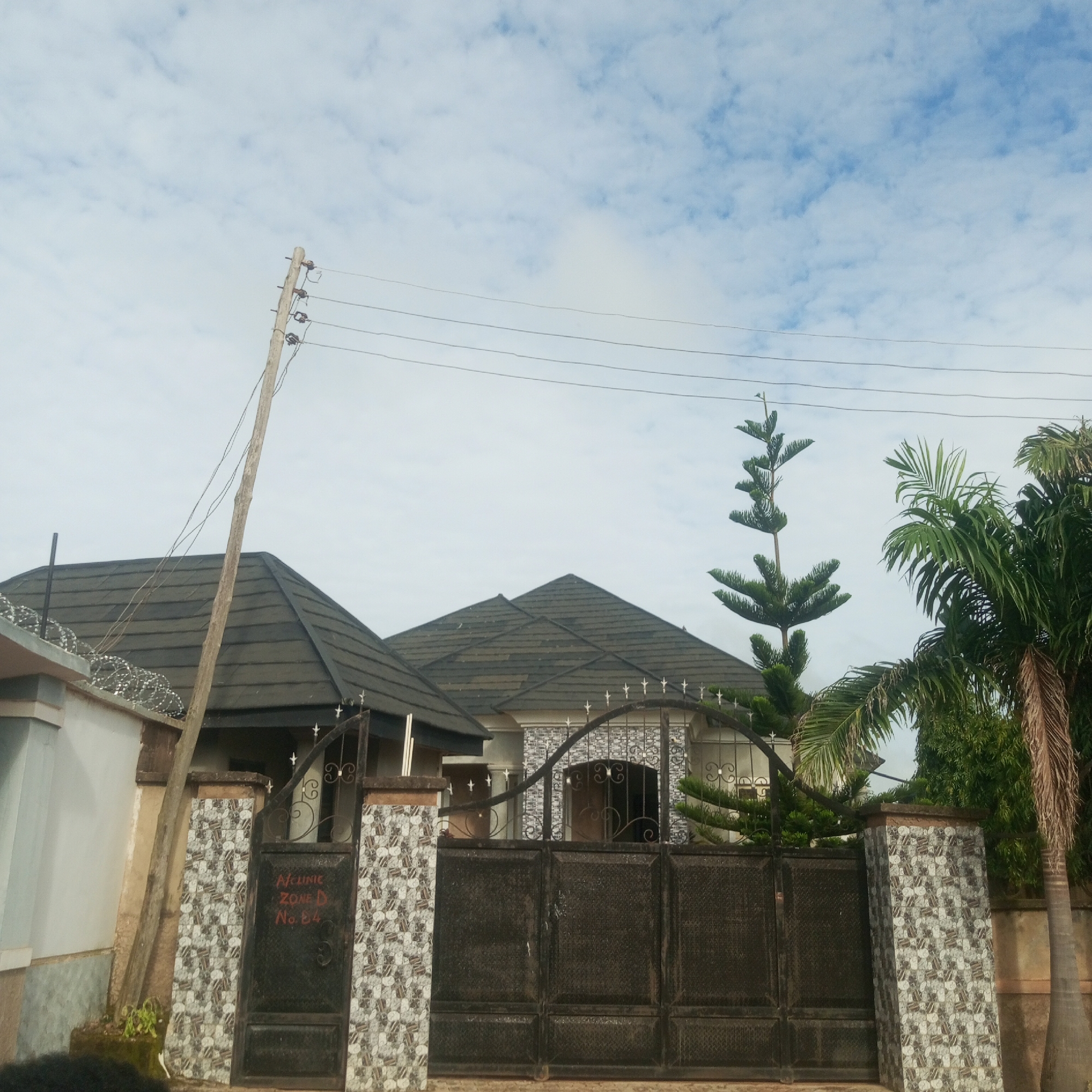 A static, eye-level, medium shot captures the exterior of a property in Jos, Nigeria. The focus is on a large, black, wrought-iron gate with decorative elements and a smaller, arched gate on the left. The gates are flanked by decorative pillars, some covered in mosaic tiles. To the left of the gate, a white wall topped with barbed wire is visible. Behind the gate, a section of a house with multiple gabled roofs covered in dark shingles is partially obscured by the gates. Some decorative tiling adorns the house facade. A tall, slender evergreen tree stands to the right of the gate, with a palm tree and other greenery behind it. Power lines are strung across the sky, supported by a wooden utility pole on the left side of the frame. The sky is overcast with scattered clouds. Red markings are visible on the left gate, reading "Alchinic ZONE D No. B4". No people are prominently featured or engaged in any discernible action.