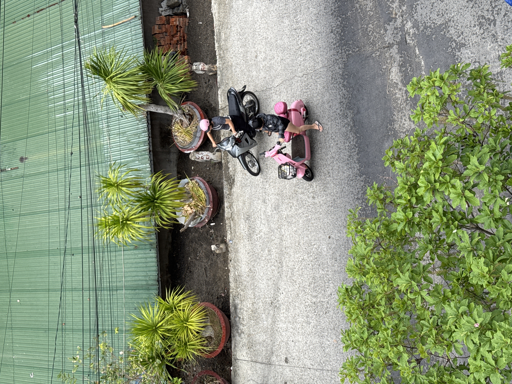 Aerial view of a street in Nha Trang Ward, Vietnam. A green corrugated metal building lines one side of the street with potted plants along the edge. A concrete pavement extends alongside the building. Two individuals stand beside two parked motorbikes, one silver and one pink. One person wears a pink helmet. A large green tree is on the opposite side of the street.