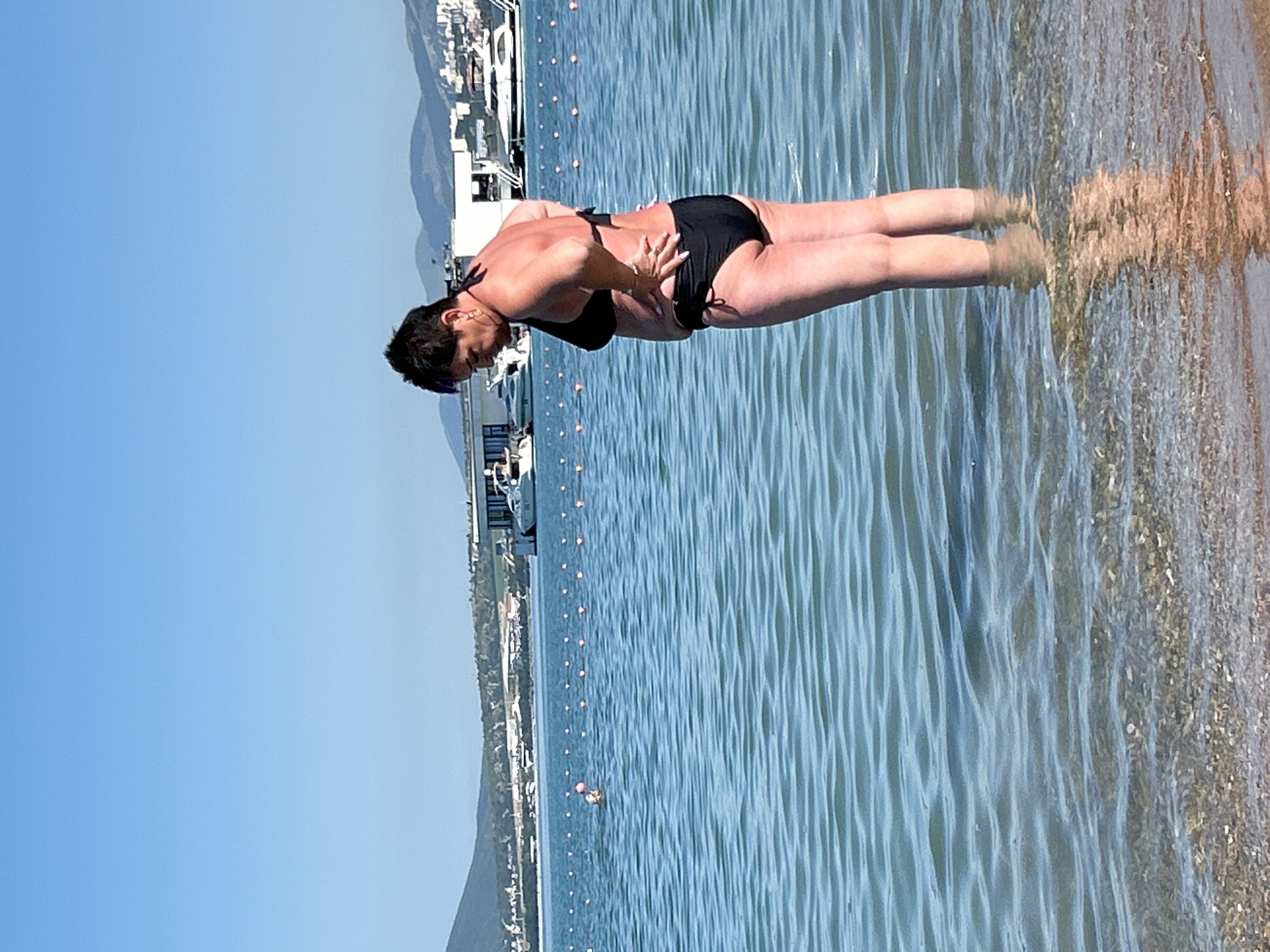 An adult, Caucasian female is standing in shallow, clear blue water near a beach, wearing a black bikini. She has short, dark hair and is facing away from the camera, with her hands on her hips. The water has gentle ripples, and the seabed is visible beneath the surface. In the background, several white yachts are anchored in the sea, and a coastal town with buildings and hills is visible on the horizon. The sky is clear and blue. The location is the city of Gelendzhik, Russia.