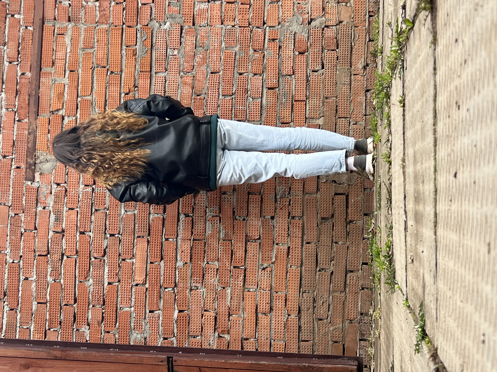 A person stands against a red brick wall, likely outdoors given the concrete sidewalk with weeds growing in its cracks to their right. The individual is wearing light-colored denim pants, dark sneakers, and a black leather jacket. They have long, wavy brown hair. A wooden structure is partially visible at the bottom of the frame. The exact location in Votkinsk, Russia cannot be determined solely from the visual information provided.