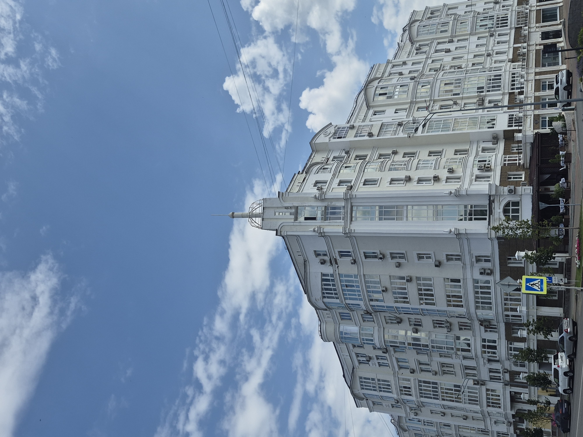 A wide-angle, slightly low-angle shot captures a modern, multi-story white apartment building against a blue sky with scattered white clouds. The building features a distinct architectural style with curved sections, numerous windows, and decorative elements. A pointed spire crowns a central section of the building. Power lines crisscross the sky above. In the foreground, a street runs along the building, with several cars parked or driving. A pedestrian crossing sign and trees line the sidewalk. The image appears to be taken in the city of Ufa, Russia, specifically on the street Lenina 134.