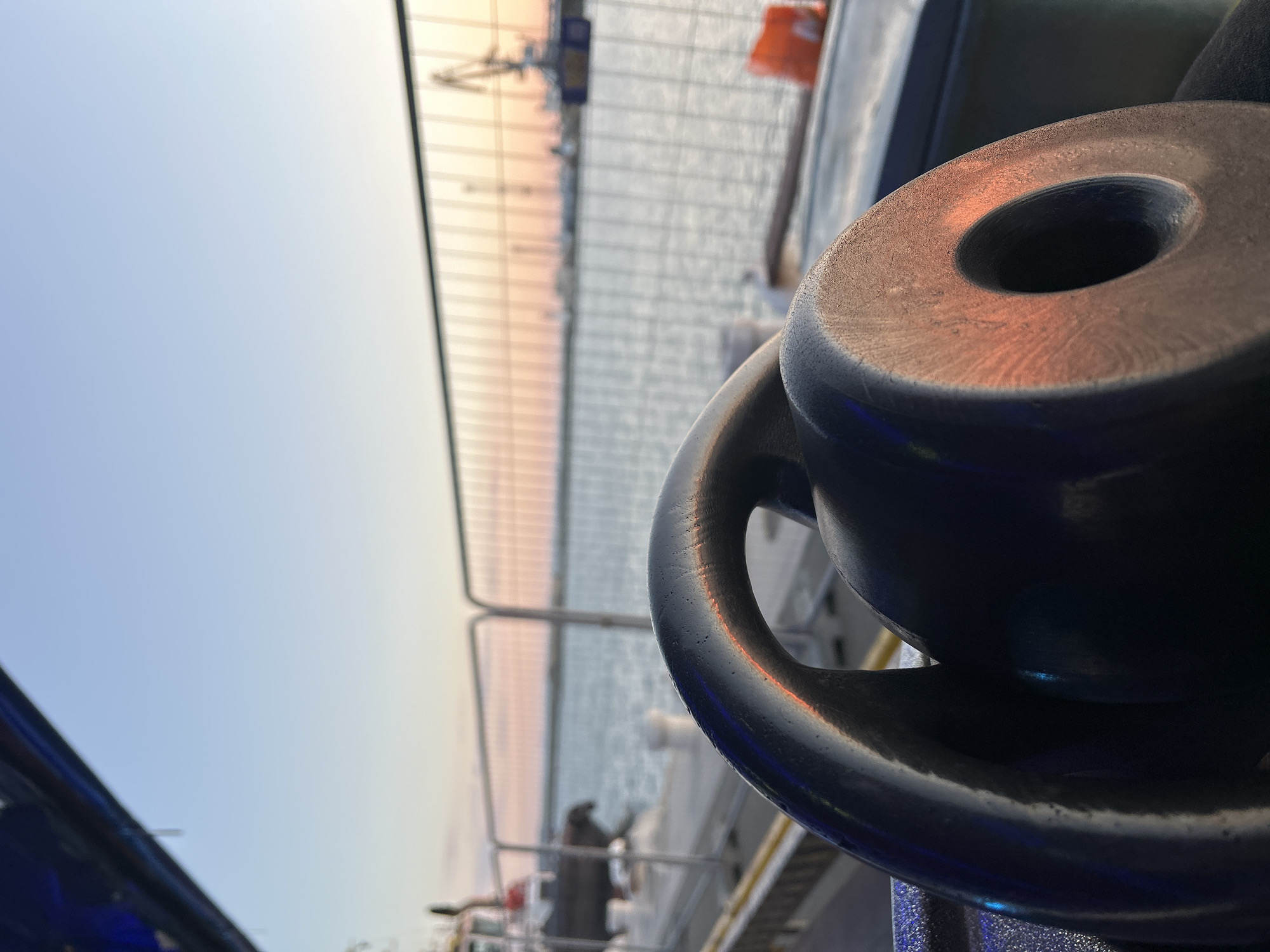 A close-up shot focuses on a dark metal mooring bollard with a prominent hole, positioned on the side of a ship or dock. In the background, a fenced waterfront area with calm water suggests a coastal location. The soft sky indicates either sunrise or sunset. The scene is consistent with Burgas, Bulgaria, due to its port and coastal setting.