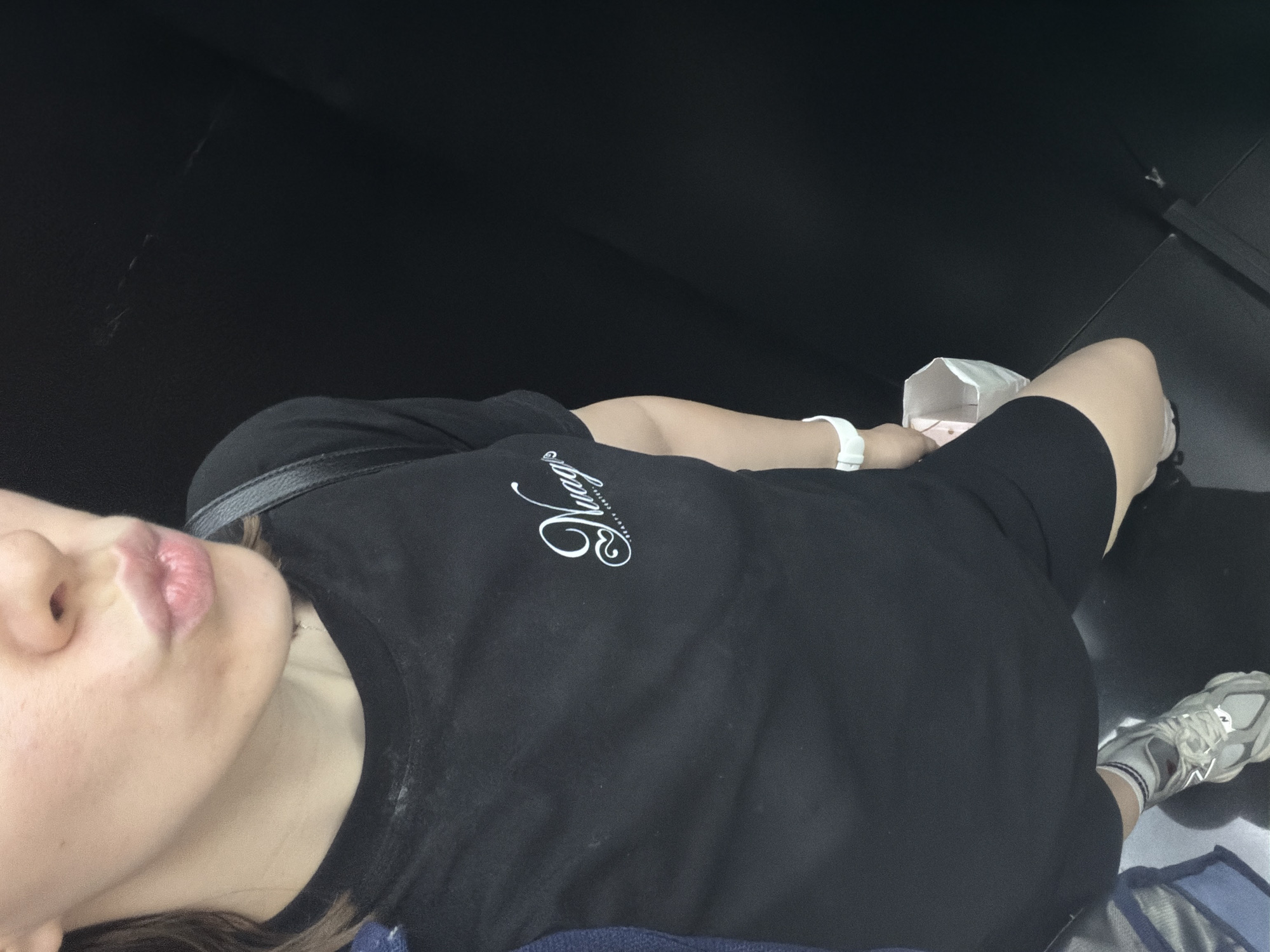 The image shows a person lying on a dark, possibly upholstered surface, with their head to the left. They are wearing a black t-shirt with white script text, the word "Nugget" is visible. The person is also wearing black shorts and athletic shoes. A white bracelet is visible on their wrist. A small, white paper container is resting on their lap. The lighting suggests an indoor setting, possibly at night or in a dimly lit area. The overall mood is relaxed.