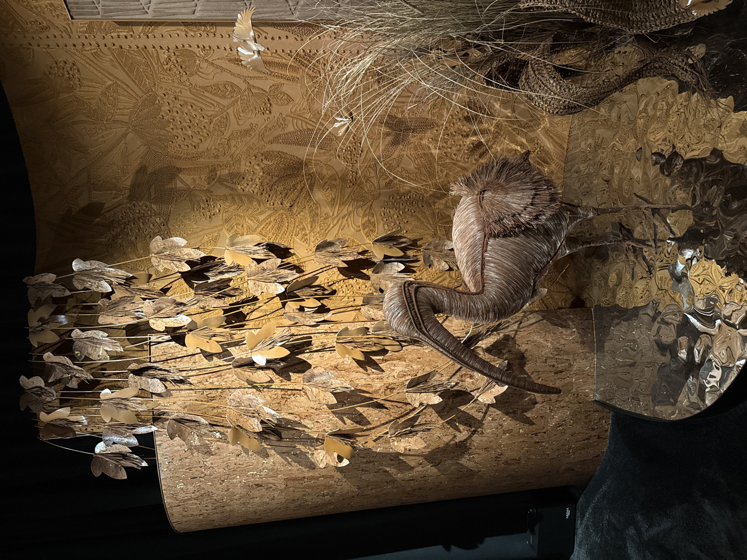 This image captures a sophisticated art installation, likely displayed indoors in a gallery or high-end retail environment in Milan, Italy.
The central subject is a striking, abstract sculpture resembling an animal, possibly a mythical creature or a stylized deer. It is crafted from tightly woven or layered natural fibers, such as raffia or twisted rope, in tones of brown and taupe, and features a textured, spiky "mane" or headpiece. The sculpture is posed dynamically, suggesting movement, and rests on a textured, possibly cork-covered base.
Behind the main sculpture, a large wall is adorned with an intricate, embossed or etched pattern of stylized leaves, vines, and possibly berries, rendered in a warm, golden-brown material that appears to be leather or a luxurious fabric. To the left, a graceful garland composed of numerous small, metallic, silver-toned leaf cutouts extends horizontally, casting dramatic shadows on the patterned wall.
To the right of the sculpture, an uneven, highly reflective surface, possibly polished metal or a distorted mirror, creates abstract reflections of the surrounding elements, adding a modern and surreal dimension to the display. Delicate, wispy strands resembling dried grass or fine feathers are visible in the upper right. Additionally, small, white, stylized bird figures appear to be in flight in the upper left and right portions of the background.
The lighting is theatrical, using spotlights to highlight the textures, metallic finishes, and intricate details, creating strong contrasts and deep shadows that enhance the three-dimensional quality of the artwork. The overall aesthetic is organic, luxurious, and naturalistic. No text is visible in the image.