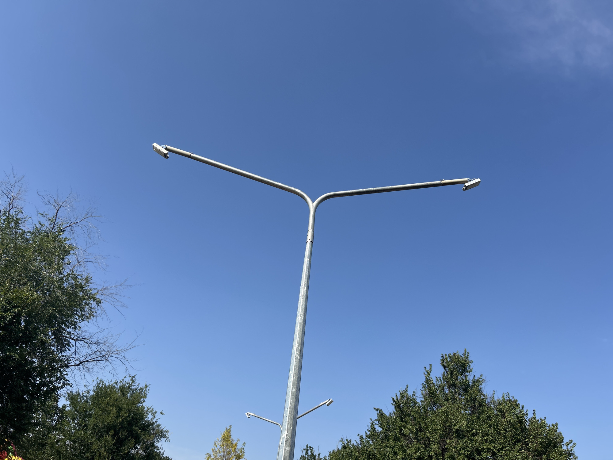 A tall, galvanized metal streetlamp with a bifurcated arm extends upwards against a clear blue sky. The streetlamp is positioned in the foreground, with its metallic surface showing some signs of weathering. Two rectangular light fixtures are attached to the ends of the arm, each angled slightly downwards. In the lower portion of the frame, lush green trees with full foliage partially obscure the base of the streetlamp. Some bare, skeletal branches of a deciduous tree are visible on the left side of the image, contrasting with the vibrant green of the other trees. The scene appears to be set in an outdoor urban environment within the city of Pivdennoukrainsk, Ukraine, likely in a park or along a street where street lighting is installed.