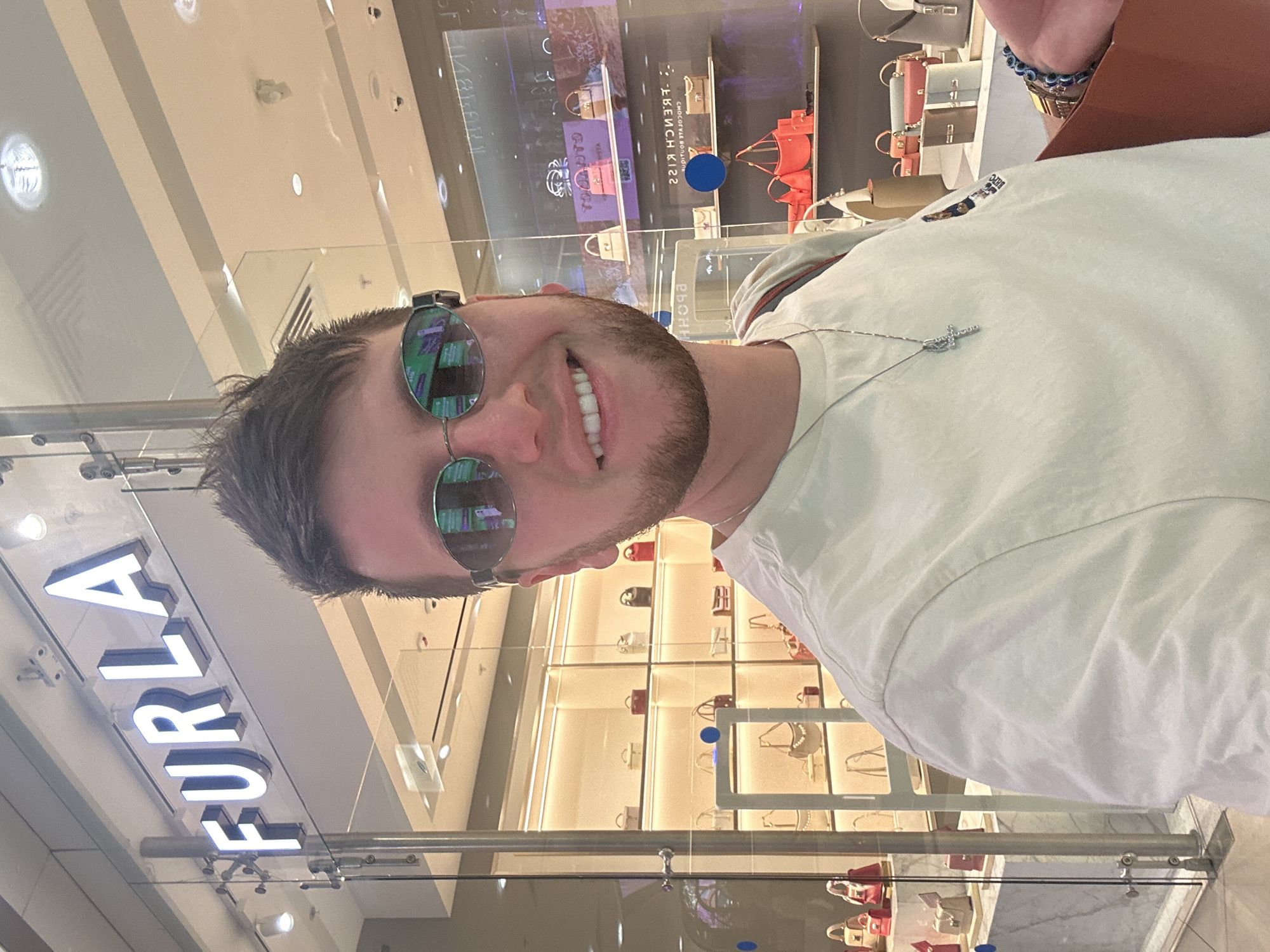 A man takes a selfie in front of a store. The store's sign reads "FURLA." The man is wearing sunglasses, a white t-shirt, and a necklace. Behind him, the store window displays various handbags on shelves. The store appears to be in a shopping mall, with reflective surfaces and bright lighting.