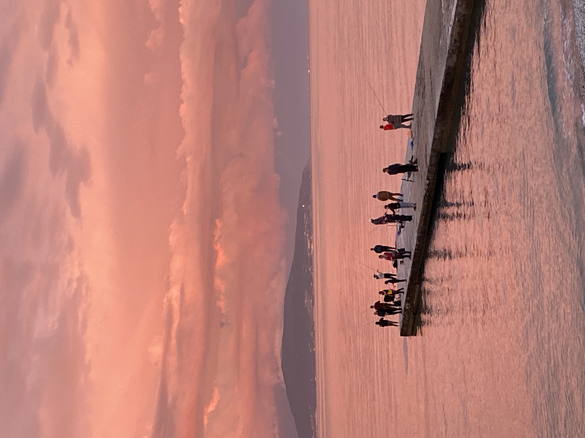 The image captures a serene coastal scene in Kabardinka, Russia, likely during either sunrise or sunset, given the striking warm light. The sky is dominated by soft pink, orange, and purple hues, with large, diffused clouds reflecting the dramatic light. These same vibrant colors are mirrored on the surface of the calm sea, creating a harmonious and tranquil atmosphere.
A long, concrete pier or breakwater extends from the right into the middle of the frame, serving as a platform for a group of approximately 10-12 people. These individuals, mostly appearing in silhouette against the reflective water, are engaged in fishing. Several are visible holding fishing rods, with lines cast into the placid water. Their postures suggest a relaxed and leisurely activity.
In the background, a distant coastline or mountainous terrain can be faintly discerned under the pastel sky. The water is exceptionally still, enhancing the reflection of the sky and light. There is no visible text in the image. The overall impression is one of peaceful recreation and the breathtaking beauty of nature at a transitional time of day on the Black Sea coast.