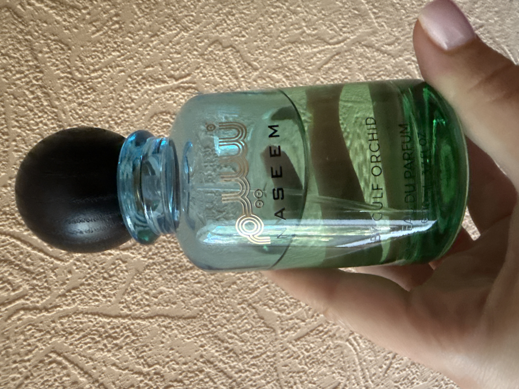 The image shows a person's hand holding a bottle of NASEEM GULF ORCHID EAU DE PARFUM. The bottle is made of green glass and has a black spherical cap. The background is a textured beige wall. The scene is indoors, and the lighting suggests it is daytime. No specific location in Okhtyrka, Ukraine is visible or indicated.
