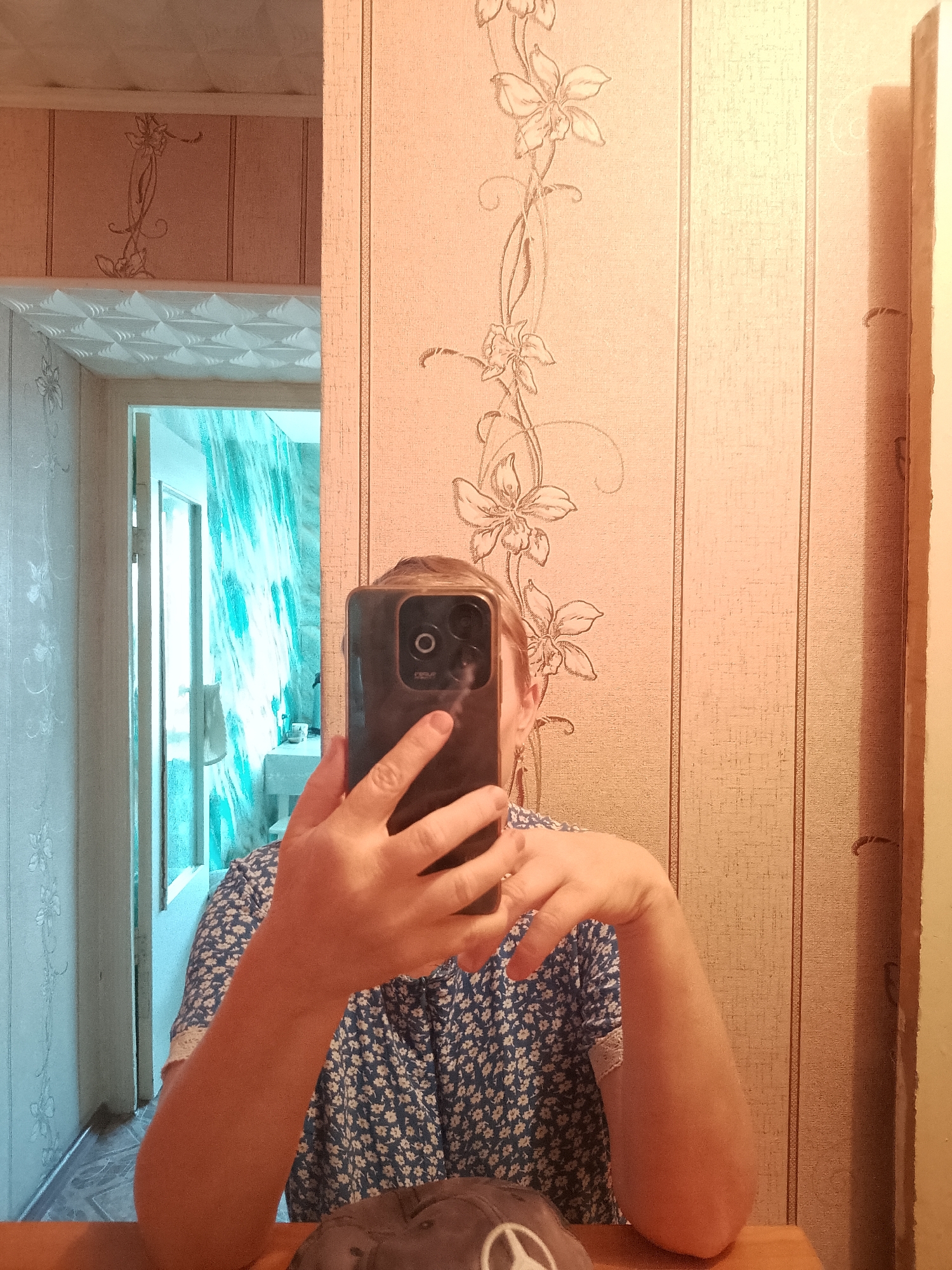 A person is taking a mirror selfie in what appears to be a room. They are holding a dark-colored smartphone in their right hand, obscuring their face. The person is wearing a blue and white floral-print shirt with white lace trim on the sleeves. Their left arm is bent, resting on their lap, and a beige bandage is visible on their left elbow. In the reflection, a doorway leads to another room with bright, abstract teal and white wallpaper. The wall behind the person in the foreground has vertical stripes and a floral vine pattern in a light peach color. A wooden surface is visible at the bottom of the frame, and a dark hat with a white emblem is resting on it. The scene is indoors, likely in a residential setting. The location in the city of Abay, Kazakhstan is not discernible from the image.