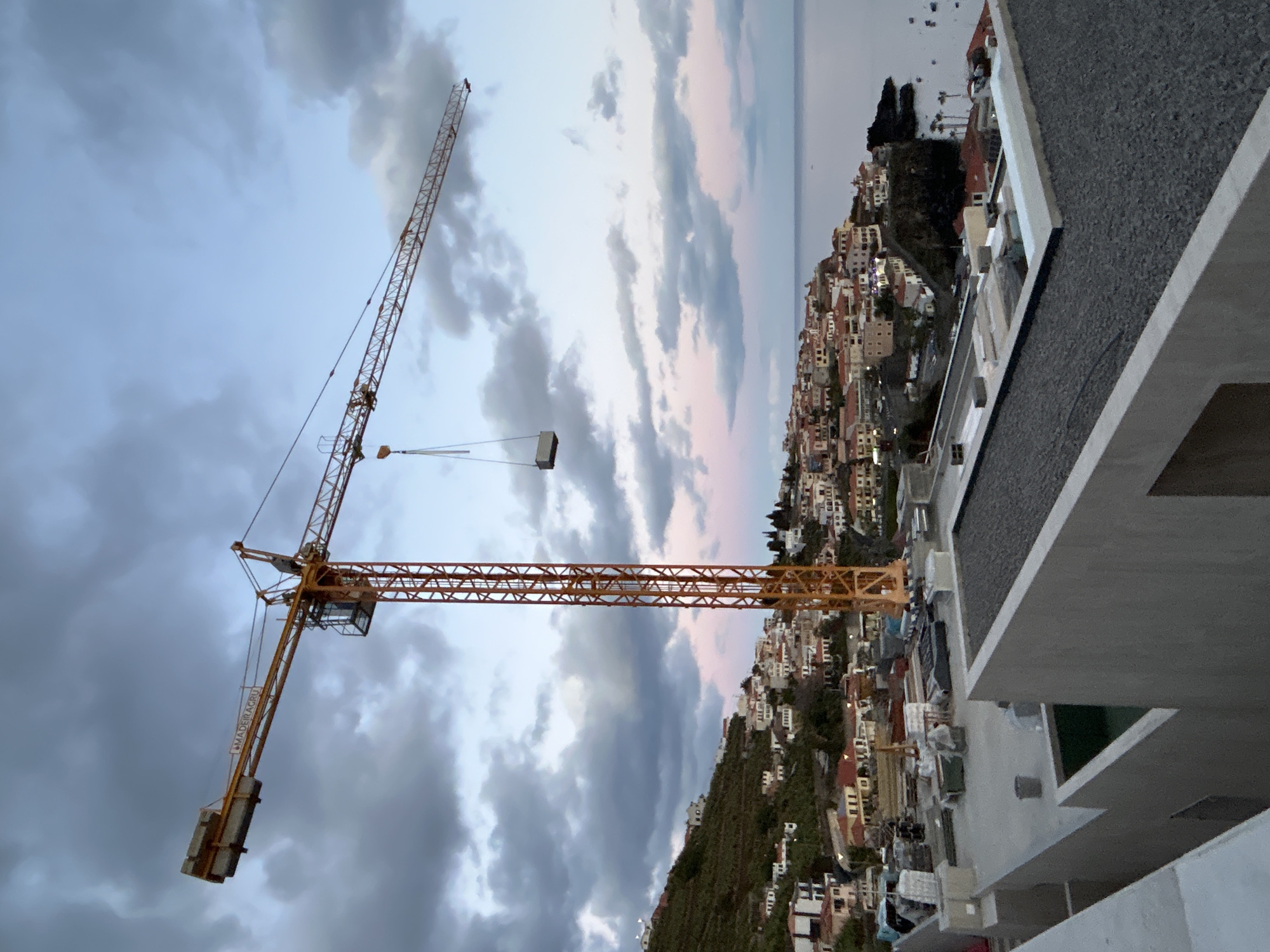 The image shows a cityscape in Câmara de Lobos, Portugal, featuring a construction crane against a sky with dramatic clouds. The crane is yellow and has a jib extending over a densely built-up area on a hillside. Buildings are predominantly white with red-tiled roofs. In the distance, the ocean meets the horizon, with a few boats visible. In the foreground is a building with a flat, dark gray roof and light gray concrete structures.