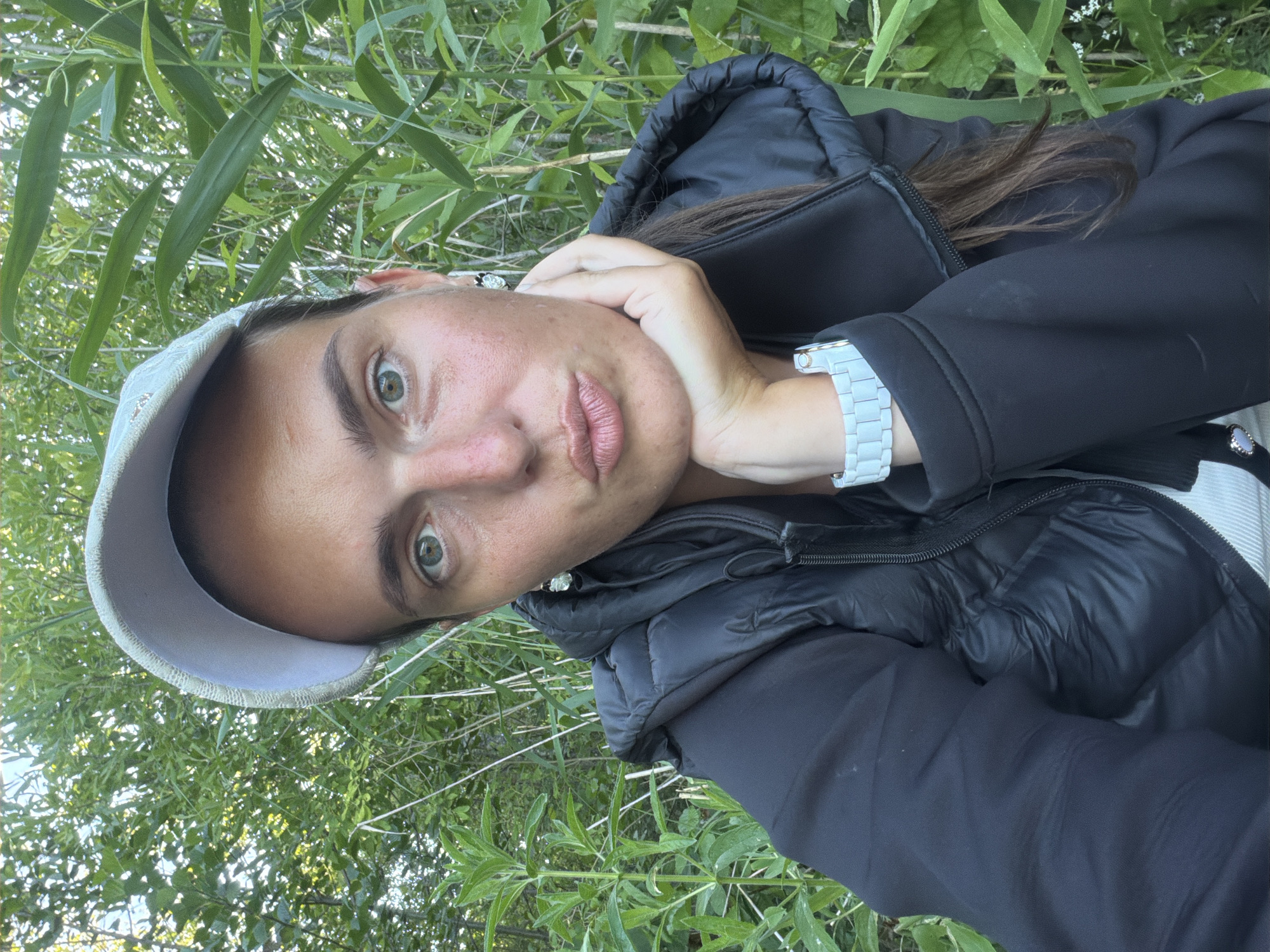 The image is a selfie taken outdoors, likely in a park or natural setting, as indicated by the dense green foliage and tall reeds in the background. The subject is a woman wearing a light-colored baseball cap, a black puffer vest over a dark jacket, and a white watch. She has her hand resting on her chin and is puckering her lips, creating a playful expression. Her eyes are open and looking towards the camera. The lighting suggests it is daytime. There is no indication of the specific location in Riga, Latvia.