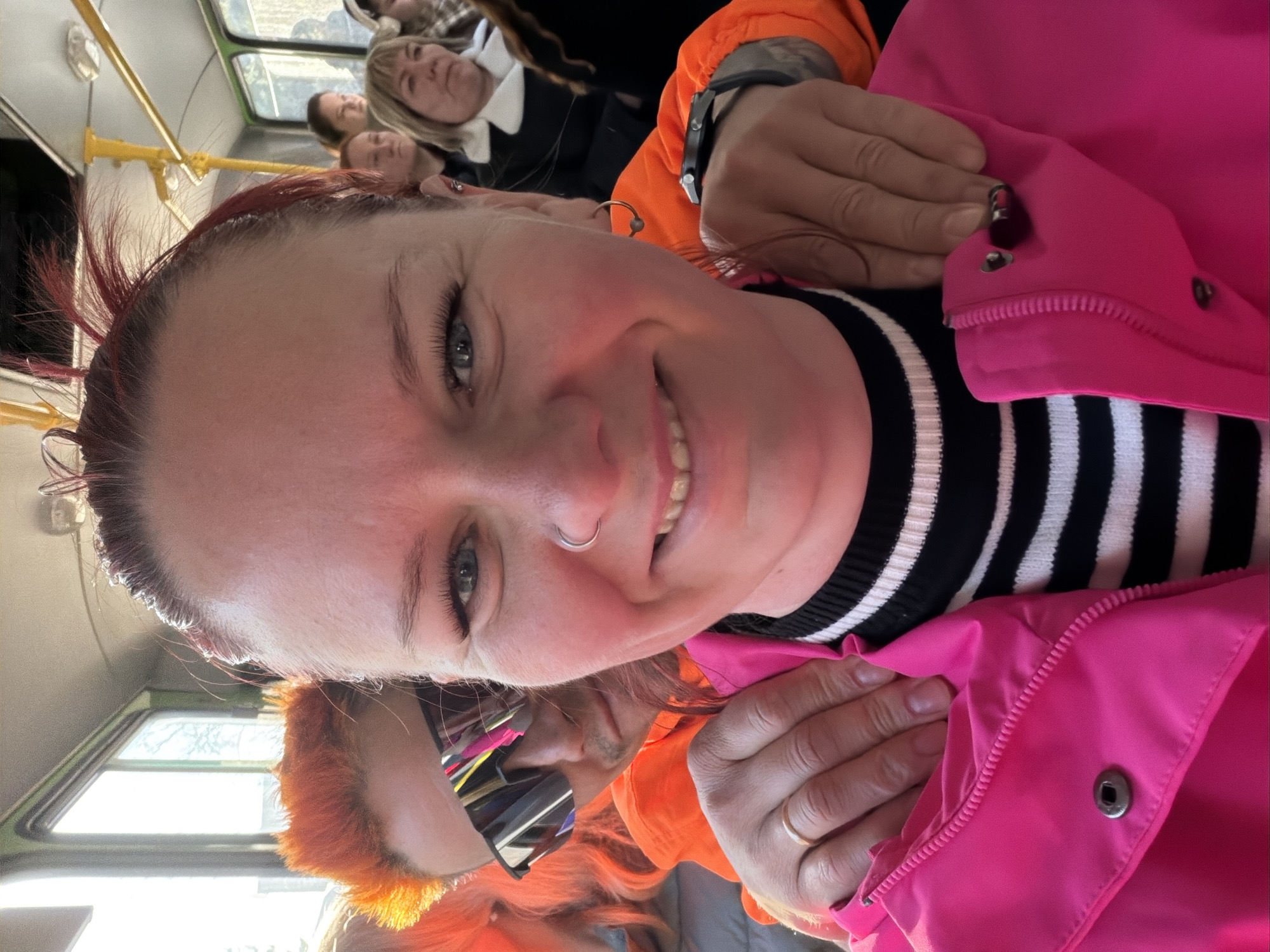 The image shows a woman taking a selfie on what appears to be a public bus. She has red hair, a nose ring, and is wearing a black and white striped shirt with a bright pink jacket. There are other people on the bus in the background. The location is Chaikovsky, Russia.