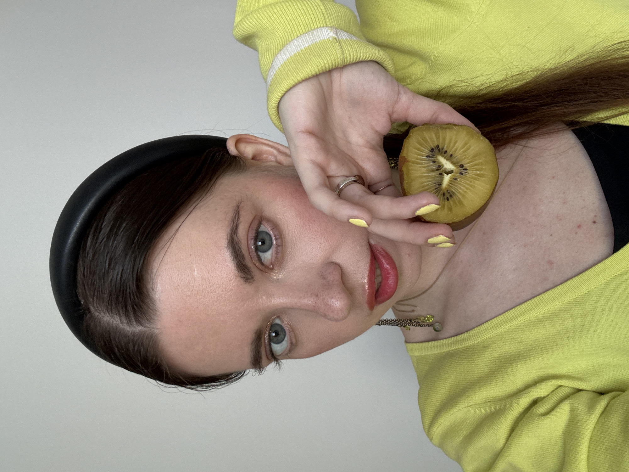 A woman with fair skin, light blue eyes, and dark brown hair is positioned in front of a plain white background. She is wearing a yellow long-sleeved top, a black tank top underneath, and a thick black headband. She is holding a slice of golden kiwi fruit in her right hand, near her face. She has a silver ring on her finger and her fingernails are painted yellow. A gold necklace is visible around her neck. The image is taken from a close-up angle and appears to be shot indoors. There is no indication of the city of Moscow, Russia.
