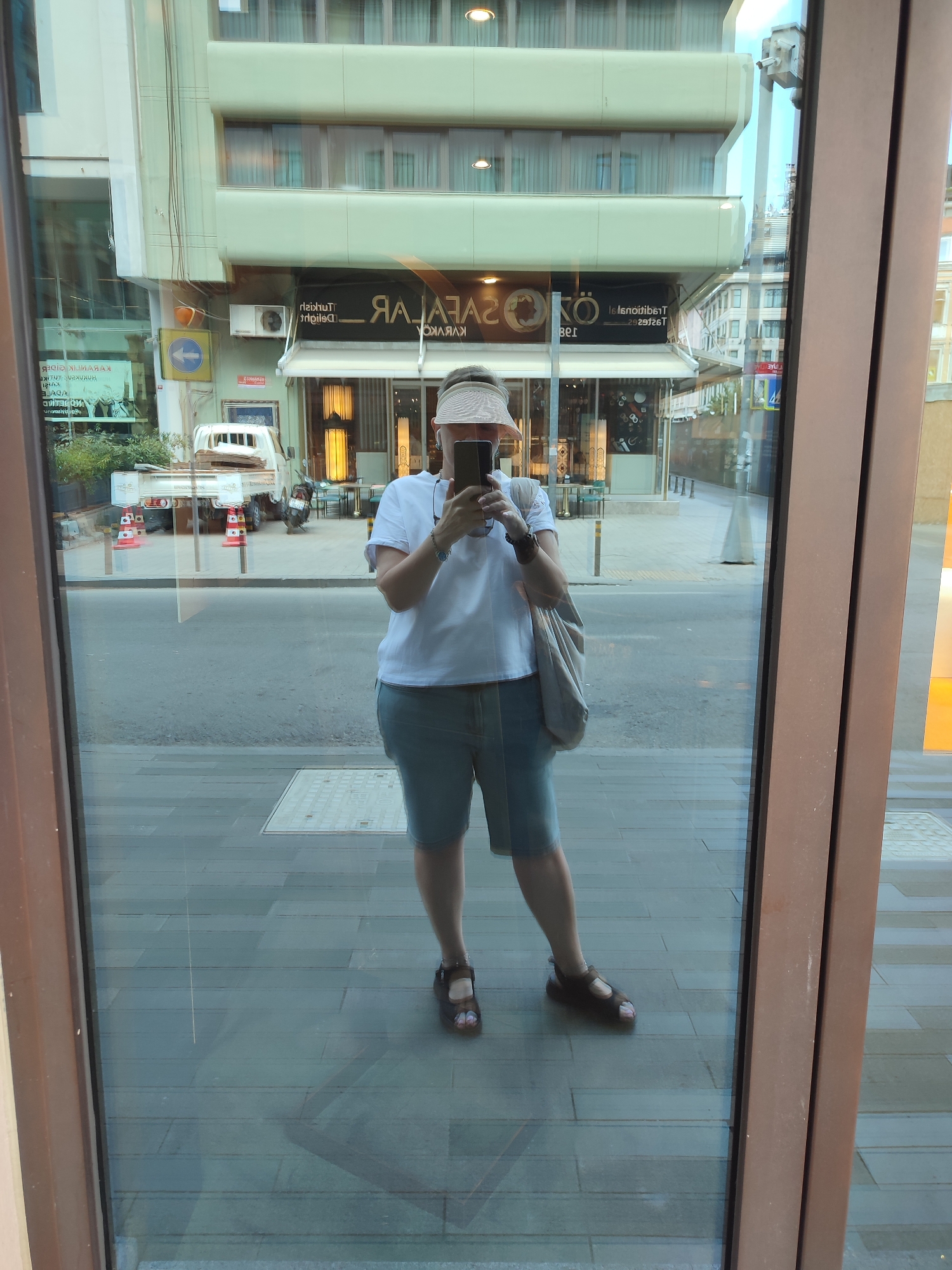 A woman is taking a selfie in a reflective window on a street in Beyoğlu, Turkey. She is wearing a white t-shirt, denim shorts, a straw hat, and dark sandals. She is holding a smartphone in her right hand and has a beige bag slung over her left shoulder. The reflection shows a street scene with buildings, trees, a white truck, and a traffic sign. One of the buildings has a sign that reads "SARAYKÖY" and "TRADITIONAL TURKISH DELIGHT". The street is paved with gray tiles.