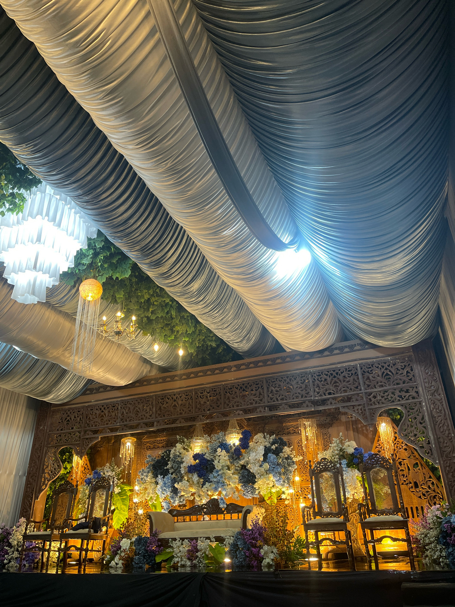 This image depicts a meticulously decorated outdoor event space, likely a wedding venue, in Bagusan, Indonesia. The scene is dominated by draped fabric ceilings, creating a sense of grandeur and enclosure. Beneath the fabric canopy, a stage is set with ornate wooden furniture, including two intricately carved chairs and a plush sofa. The stage is adorned with abundant floral arrangements in shades of blue, white, and purple, interspersed with greenery. Several decorative light fixtures, including chandeliers and string lights, are strategically placed to illuminate the space. The overall ambiance is one of elegance and festivity.