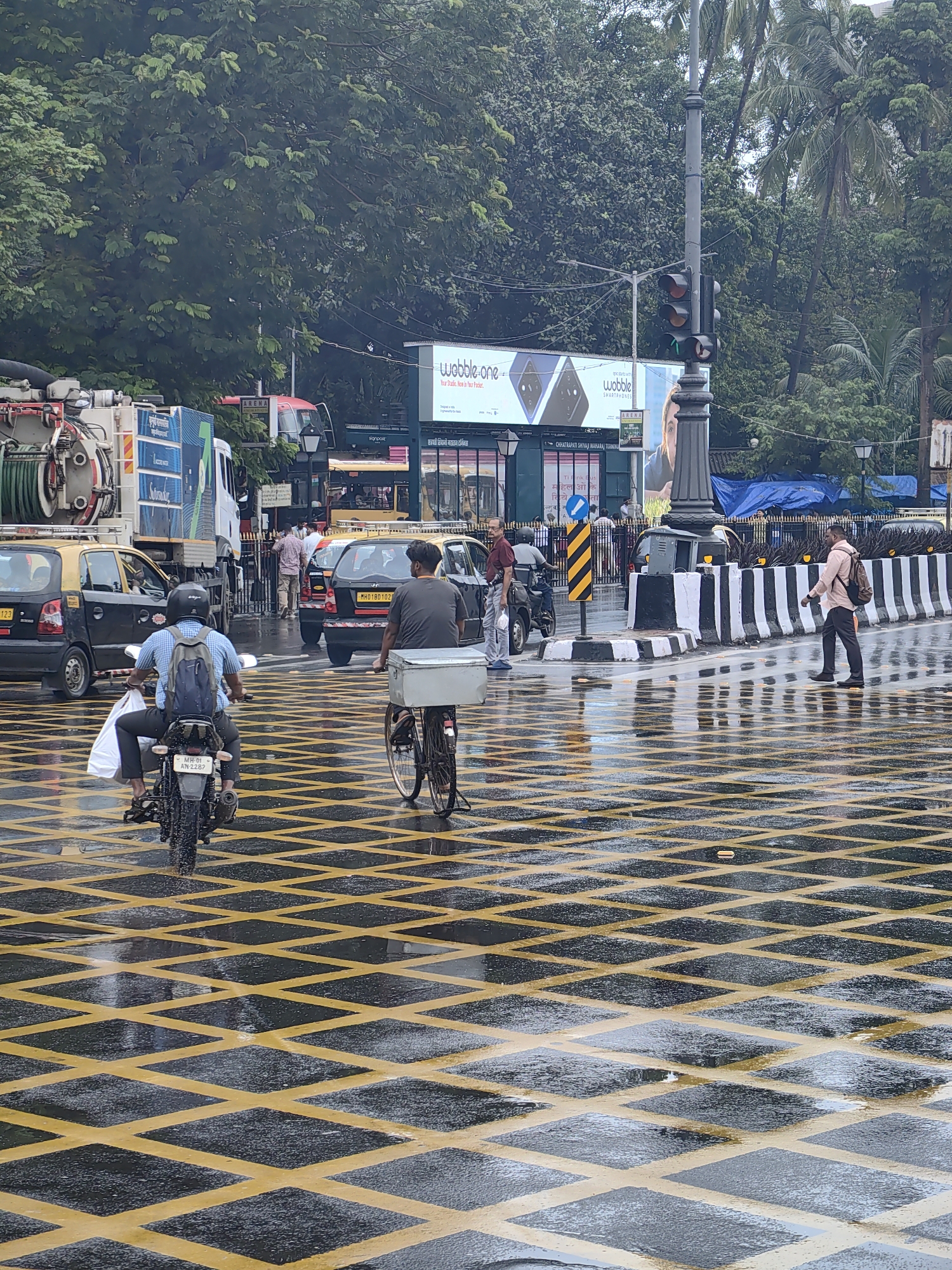 The image depicts a busy street intersection in Mumbai, India, on a rainy day. The road surface is wet and reflects the surrounding elements. Yellow diamond-shaped markings on the road indicate a no-stopping zone. Several vehicles are present, including a truck with a hose, a yellow taxi, and a motorcycle. A person on the motorcycle is wearing a blue checkered shirt and a backpack, carrying a white bag. To the right, a person is riding a bicycle with a metal box attached to the front. In the background, there are trees, a traffic light, and billboards. A pedestrian in a light pink shirt is walking away from the camera. The overall scene captures the hustle and bustle of urban Indian life, even amidst the rain.