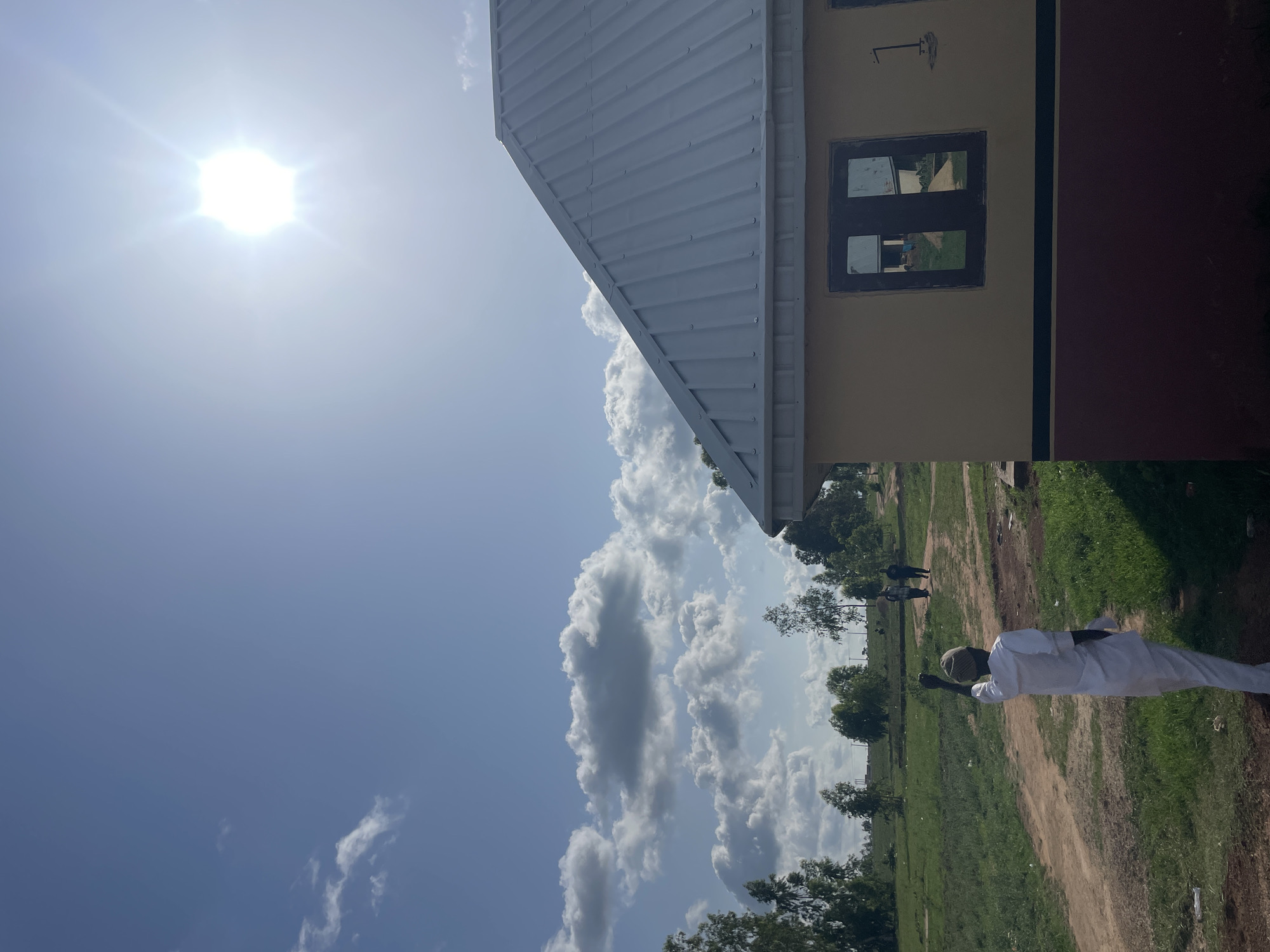 The image shows an outdoor scene in Durumi, Nigeria. In the foreground, a person wearing a white garment and a cap stands on a dirt path surrounded by grass and scattered trees. The person appears to be holding something in their hand. In the background, there are more figures visible along the path, suggesting activity or movement. Part of a building is visible on the left side of the frame, featuring a white metal roof and a window. The sky is predominantly blue with scattered cumulus clouds and a bright sun. The overall environment suggests a rural or semi-rural area, with the presence of both natural elements and structures.