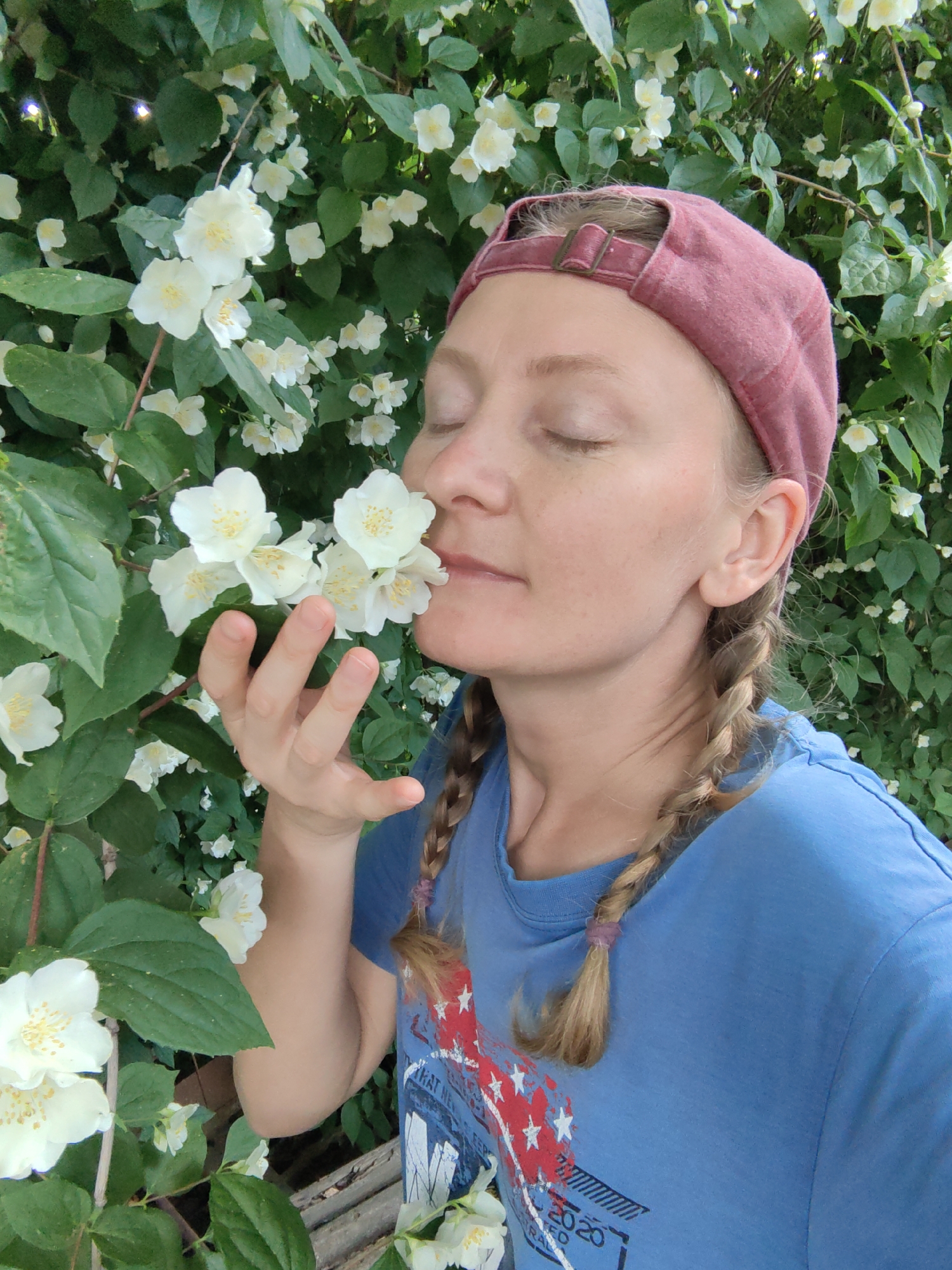 An outdoor close-up depicts a female subject in Ароматновское сельское поселение, Ukraine. She wears a faded red baseball cap reversed, secured by a bronze-colored buckle, and a blue short-sleeved t-shirt with a red and white graphic partially displaying "2020" on its left side. Light-colored hair is styled in two braids, visible over her shoulders. Her eyes are closed, and her right hand holds a cluster of white, five-petaled flowers with yellow centers to her nose. The flowers, consistent with jasmine or mock orange, are densely arrayed on a bush with dark green leaves, forming both the foreground and background of the scene.