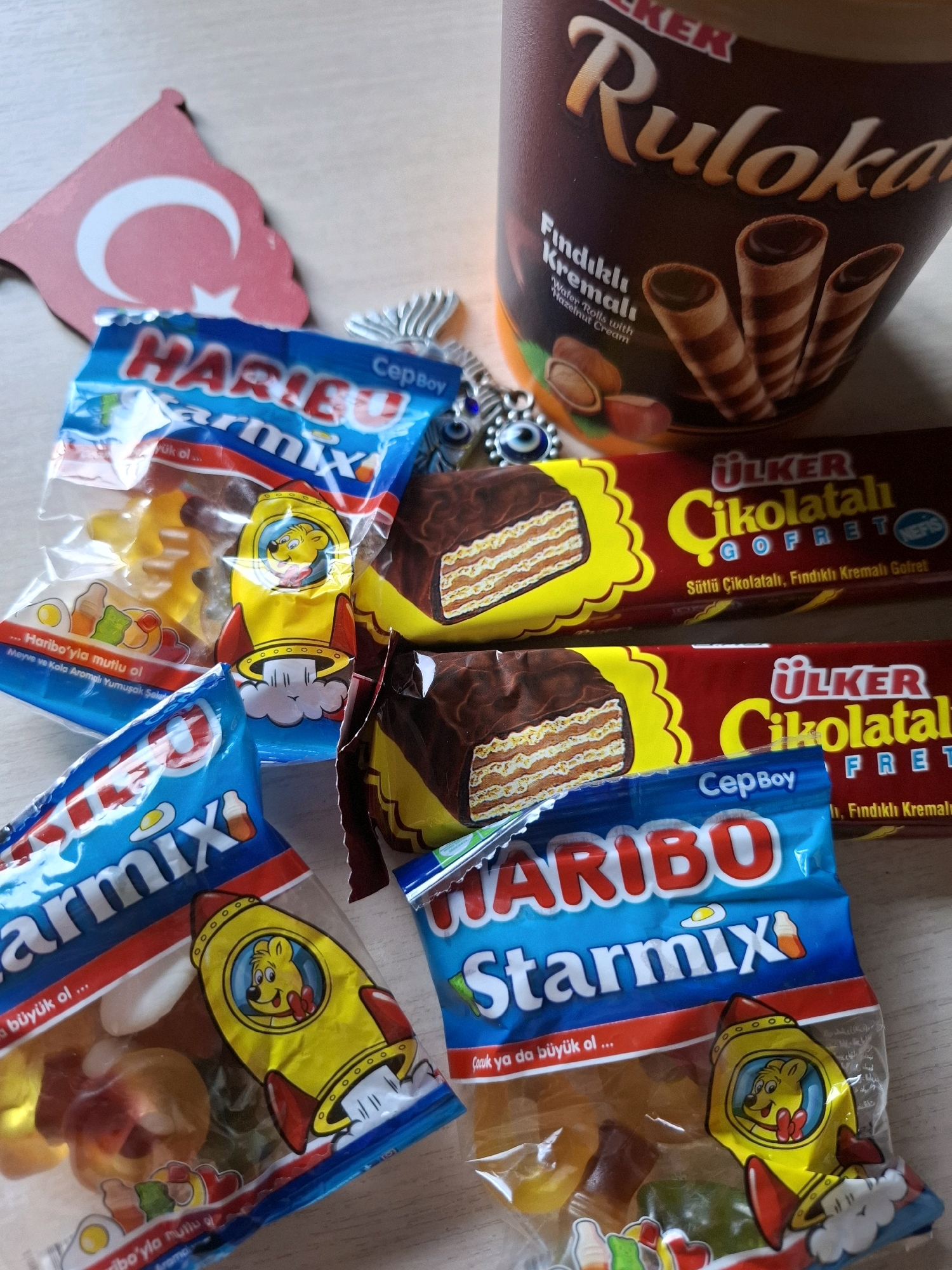 The image displays a collection of confectionery items, including several packages of Haribo Starmix gummy candies and two Ulker Cikolatalı Gofret chocolate wafers. A cylindrical container labeled "Ulker Ruloka" with wafer rolls is also visible. A small wooden Turkish flag and a decorative evil eye charm are placed amongst the snacks. The items are arranged on a light-colored, textured surface, likely a table. There are no people or actions depicted in the scene. The location is not specified as Tyumen, Russia, or any other city.