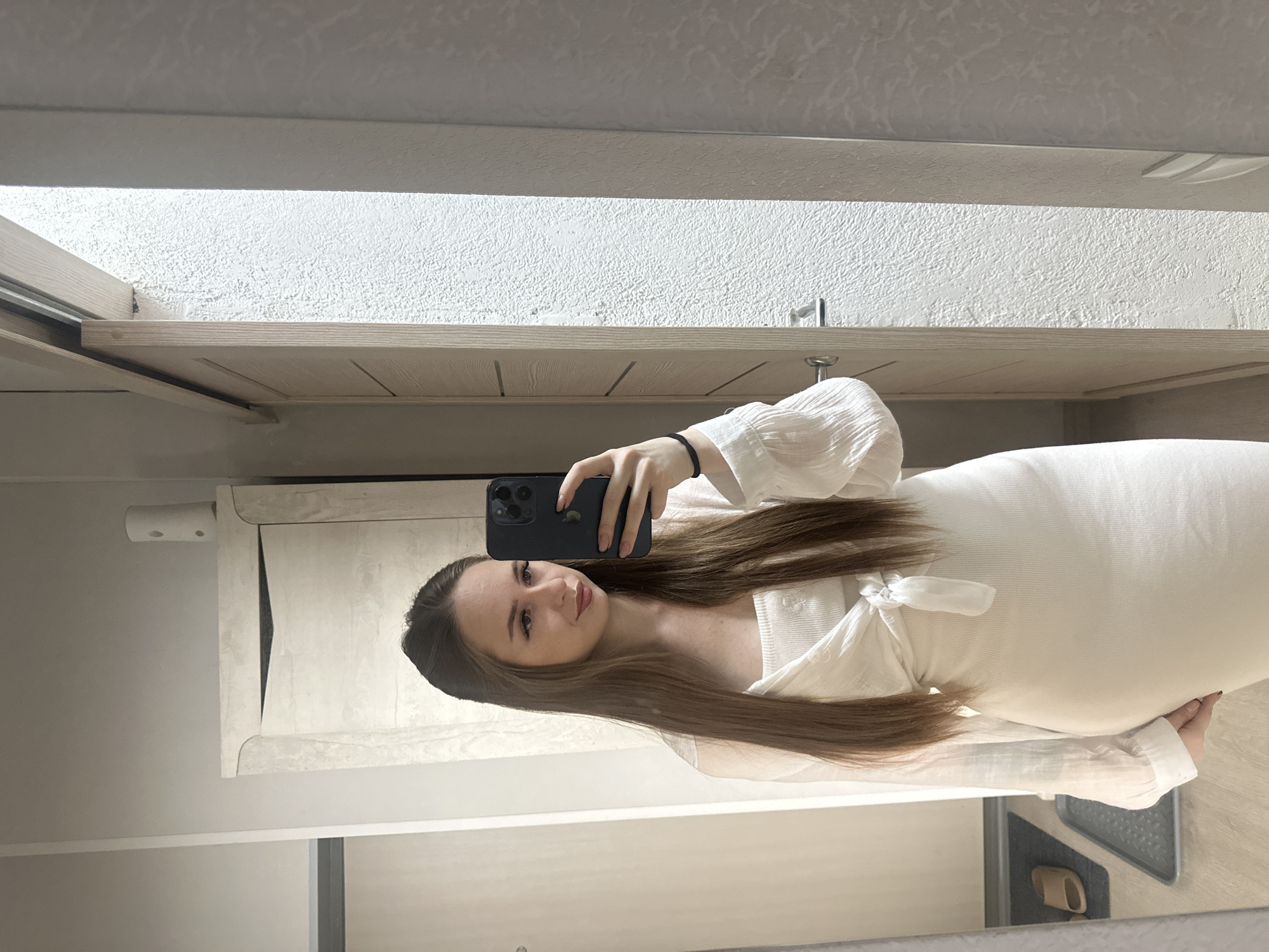 A young woman with long brown hair pulled back into a half-up style takes a selfie in a mirror. She is wearing a white ribbed tank top with a white sheer long-sleeved shirt tied at the waist. She is holding a dark-colored smartphone in her right hand. The background appears to be a hallway or dressing area with a wooden sliding door and a white textured wall. There is a tall white cabinet to the left of the frame.