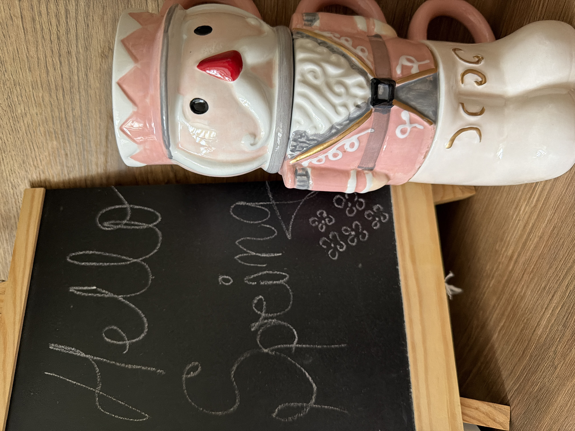 The image shows a chalkboard with "Hello Spring" written in chalk. To the right of the chalkboard is a stack of nutcracker-themed mugs, predominantly pink and white, with intricate details on the uniforms. The mugs are placed on a wooden surface. The scene appears to be indoors. There is no information about the location in Pazardzhik, Bulgaria.