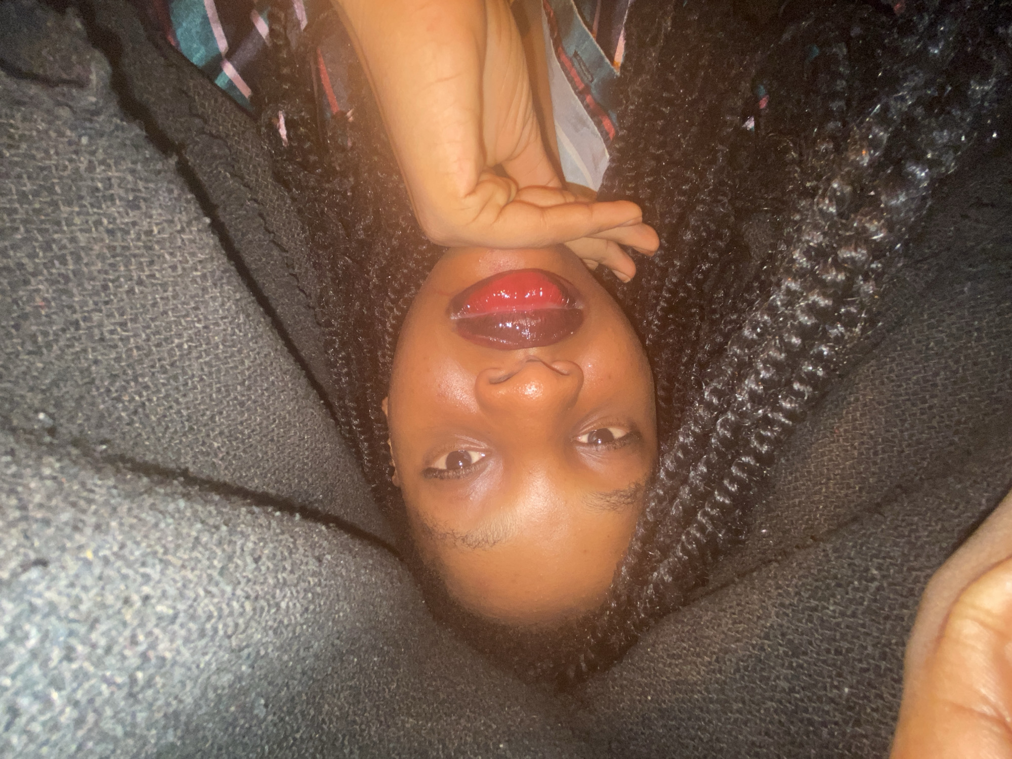 A close-up, upside-down shot depicts a person with brown skin and braided hair, positioned between what appears to be dark gray upholstered furniture, possibly a couch. The person's face is visible, with a focus on their lips, which are coated in a glossy, two-toned lipstick. One of their hands is raised near their face. The background consists of the braided hair and the upholstered surfaces.
