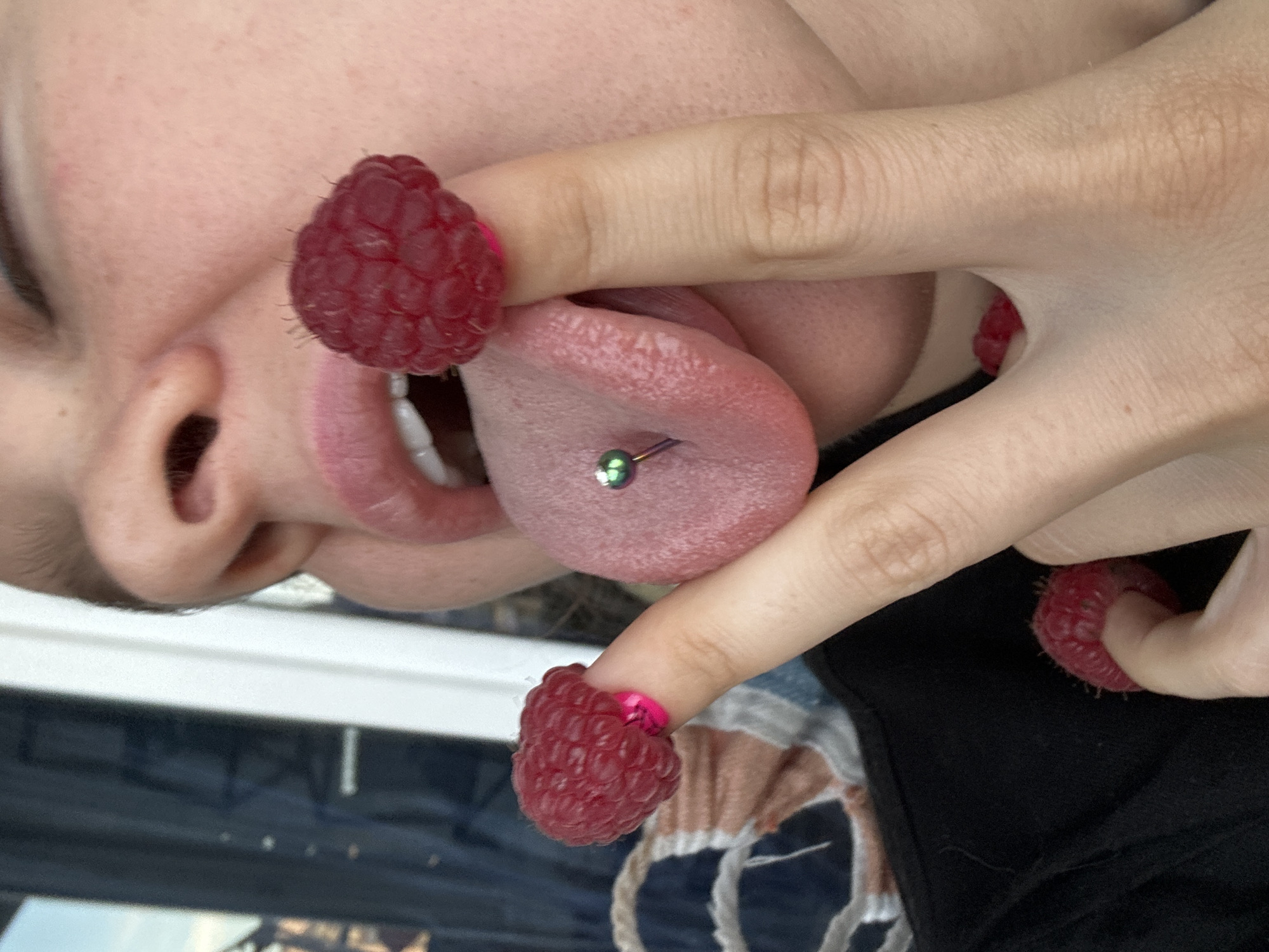A close-up, slightly skewed photograph depicts a person with a pierced tongue, using their fingers as a makeshift frame around their mouth. Raspberries are placed over the tips of their fingers. The environment is indiscernible, providing no evidence to place the image within Vladimir, Russia, or any other specific location. The person has fair skin. The tongue piercing is visible, and the person's teeth are partially visible.