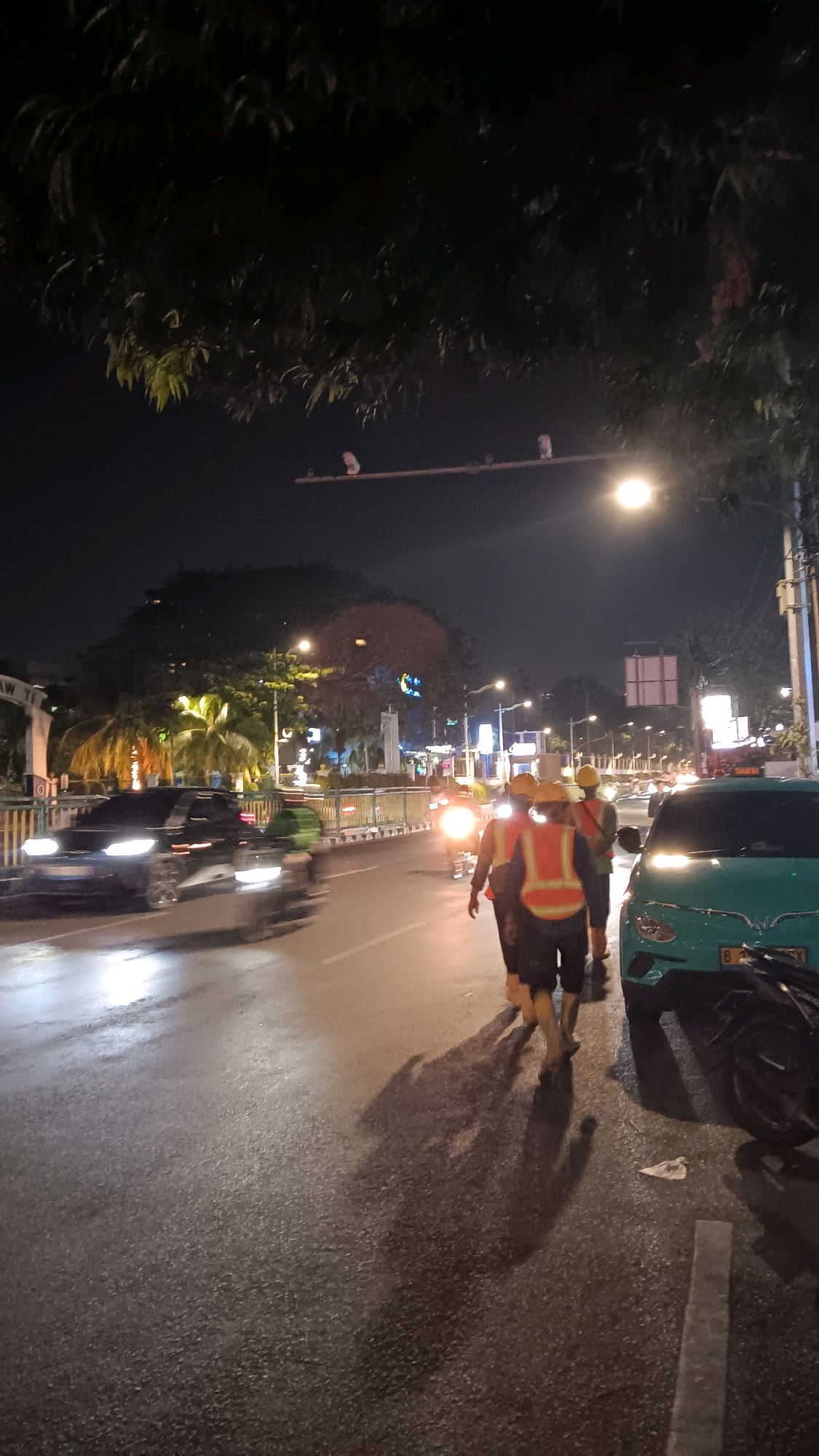 The image shows a nighttime street scene in South Jakarta, Indonesia. The street is wet, indicating recent rain or high humidity. In the foreground, three individuals wearing bright orange safety vests and yellow hard hats are walking away from the camera, their silhouettes cast as long shadows on the wet pavement. They appear to be construction workers or road maintenance personnel.
To the right, a turquoise taxi is parked, with its headlights illuminating the road. A motorcycle is also parked behind the taxi, its wheel visible. Further down the road, traffic lights and streetlights cast a warm glow, illuminating the urban environment. Cars are visible in the distance, their headlights and taillights creating streaks of light due to the motion blur. Trees line the sides of the street, their leaves dark against the night sky. The overall impression is a busy urban street at night, with ongoing activity despite the late hour.