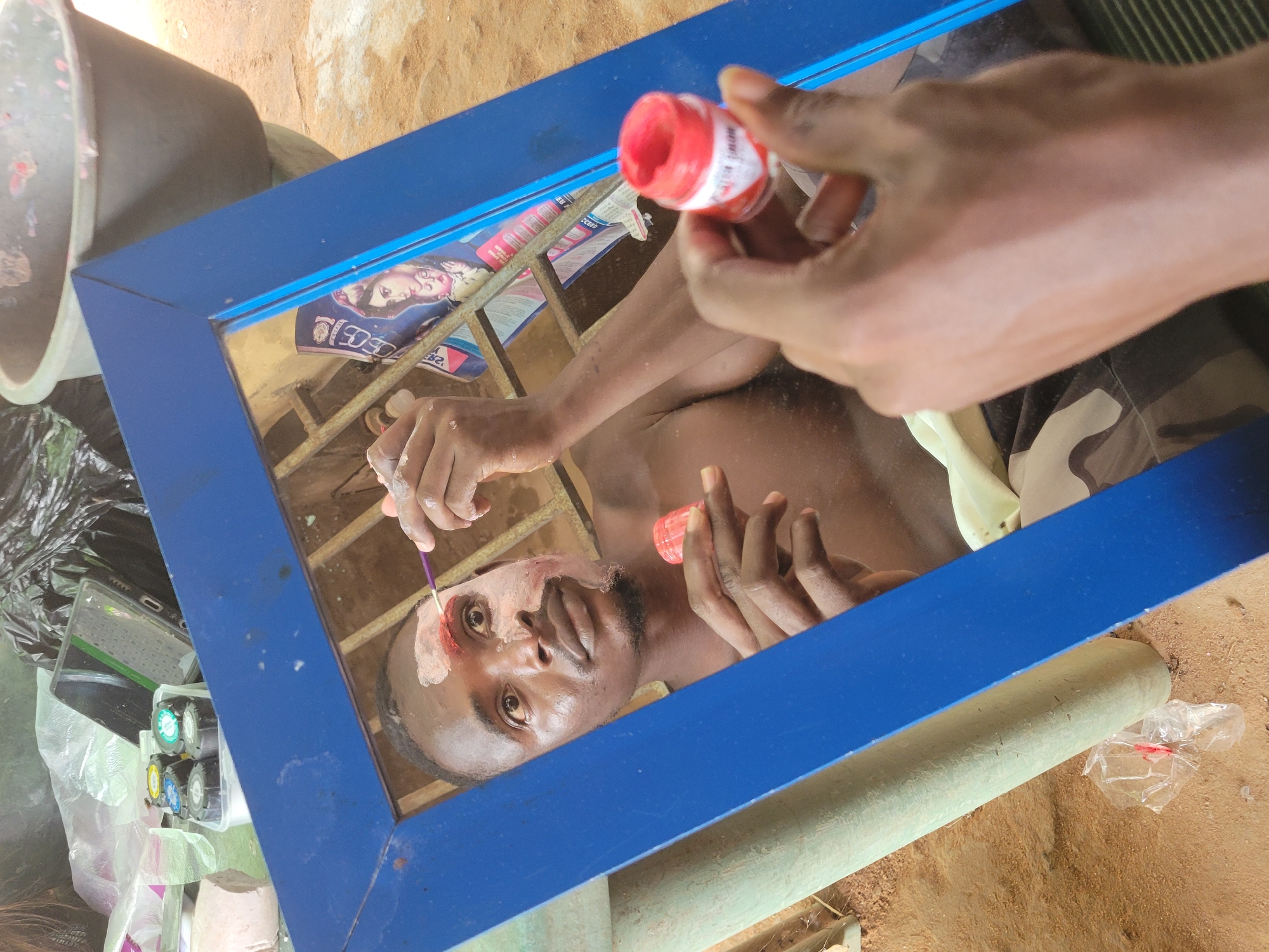 A man is applying red paint to his face with a brush, as reflected in a blue-framed mirror. He is shirtless and there is white paint already on his face. The mirror is surrounded by various items, including a black bucket, a keyboard, small containers with colorful inserts, a rolled object, and a plastic bag. The background appears to be an outdoor sandy area. The specific location within the city of Unknown, Unknown is indeterminable from the image.