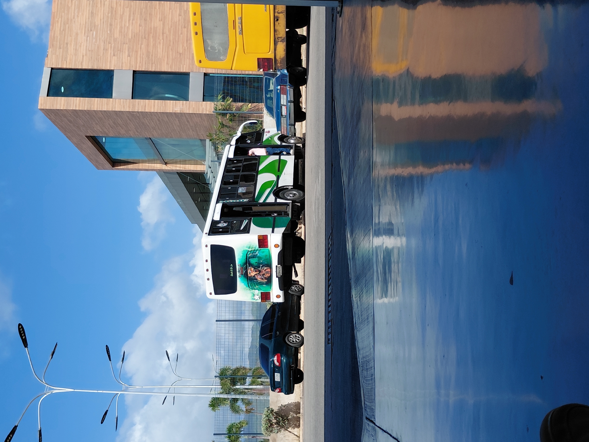 In Juan Griego, Venezuela, the image captures a street scene with a modern building in the background. A white and green bus is parked, and a person is visible near the bus door. A dark green sedan is parked to the left of the bus, and a blue sedan is positioned between the white bus and a yellow truck. The ground in the foreground is a highly reflective surface, likely wet pavement, showing reflections of the sky and surrounding structures. Tall lamp posts with multiple lights are visible on the left side of the image. Palm trees and a fence are also present in the background.