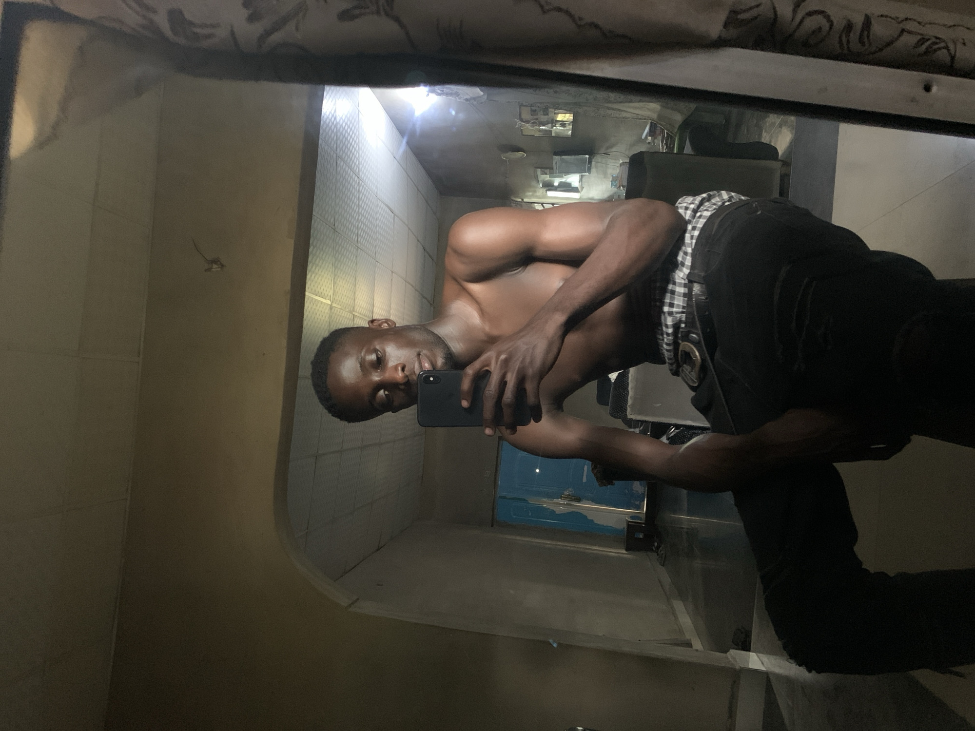 A shirtless man is taking a selfie in a mirror. He is wearing dark pants and a checkered shirt tied around his waist. The reflection shows a hallway with tiled walls and a blue door. There are some items on a shelf or table visible in the background. The location is possibly Gbeleko, Nigeria, based on the prompt.