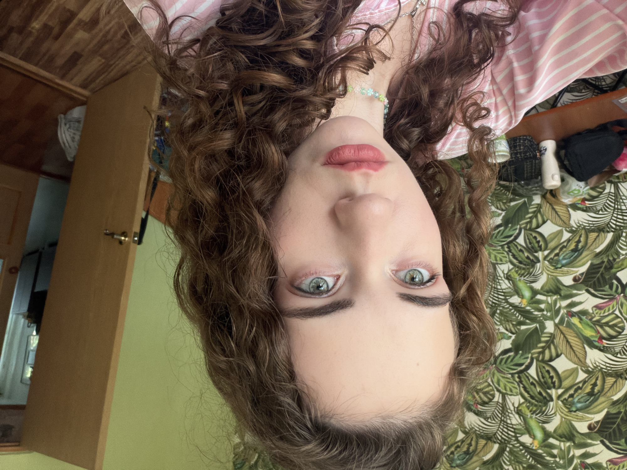 A young woman with long, curly brown hair is positioned centrally in the frame, facing the camera. Her eyes are a striking green, and she has fair skin with rosy cheeks. She is wearing a pink and white striped top and a beaded necklace. The background features a wall with a tropical-patterned wallpaper on the left and a light green wall on the right. A wooden door is slightly ajar to the right, revealing a glimpse of a room with wooden flooring. A desk with various items, including a black bag and bottles, is visible on the left side of the image. The lighting suggests an indoor setting. There is no indication of the specific location within Zavodoukovsk, Russia.