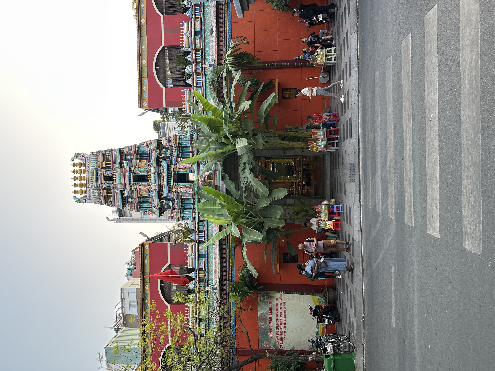 The image depicts the Mariamman Hindu Temple in Ho Chi Minh City, Vietnam. The temple features a tall, ornate gopuram (tower) adorned with colorful sculptures and intricate details. In front of the temple, banana trees grow, partially obscuring the entrance. Pedestrians and vendors are present on the sidewalk, and a crosswalk marks the street. A sign in Vietnamese, "ĐỀN BÀ MARIAMMAN," identifies the temple and indicates its status as a city-level architectural heritage site. Motorcycles and bicycles are parked nearby, adding to the busy urban environment. The background shows other buildings and a hazy sky.