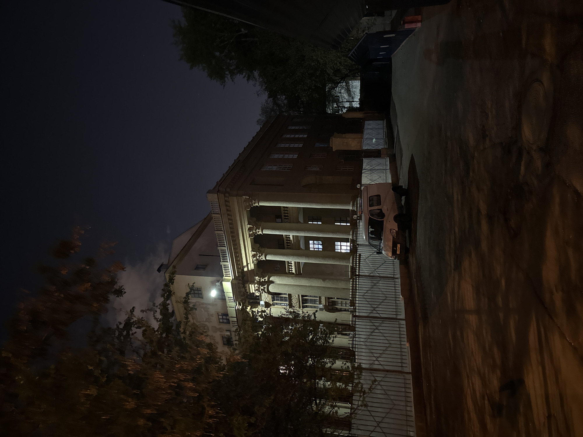 The image shows a building with a classical facade featuring columns, likely located in Moscow, Russia. The building is lit from within, illuminating its windows and columns. A vehicle is parked next to the building. A wrought iron fence runs along the side of the building, separating it from the street. Trees are present in the foreground, partially obscuring the view. The sky is dark, indicating it is nighttime. The street is paved and reflects the light from the building and street lamps.