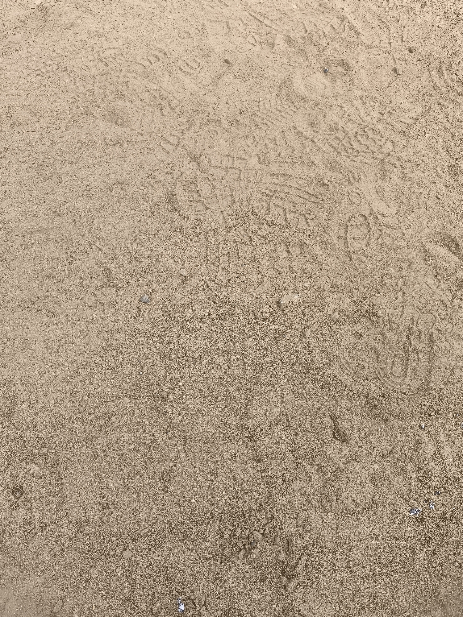 The image shows a close-up, overhead view of a sandy ground surface imprinted with numerous shoe prints. The sand is a light brown or tan color and appears to be somewhat dry, with small pebbles and debris scattered throughout. The shoe prints are varied, indicating multiple individuals have walked across the area. Some prints are clear and distinct, showing tread patterns, while others are fainter or partially obscured. The setting is a natural outdoor environment, consistent with a park, trail, or unpaved area within the village of Osieki Lęborskie, Poland. There are no people or specific objects visible beyond the ground and the imprints.