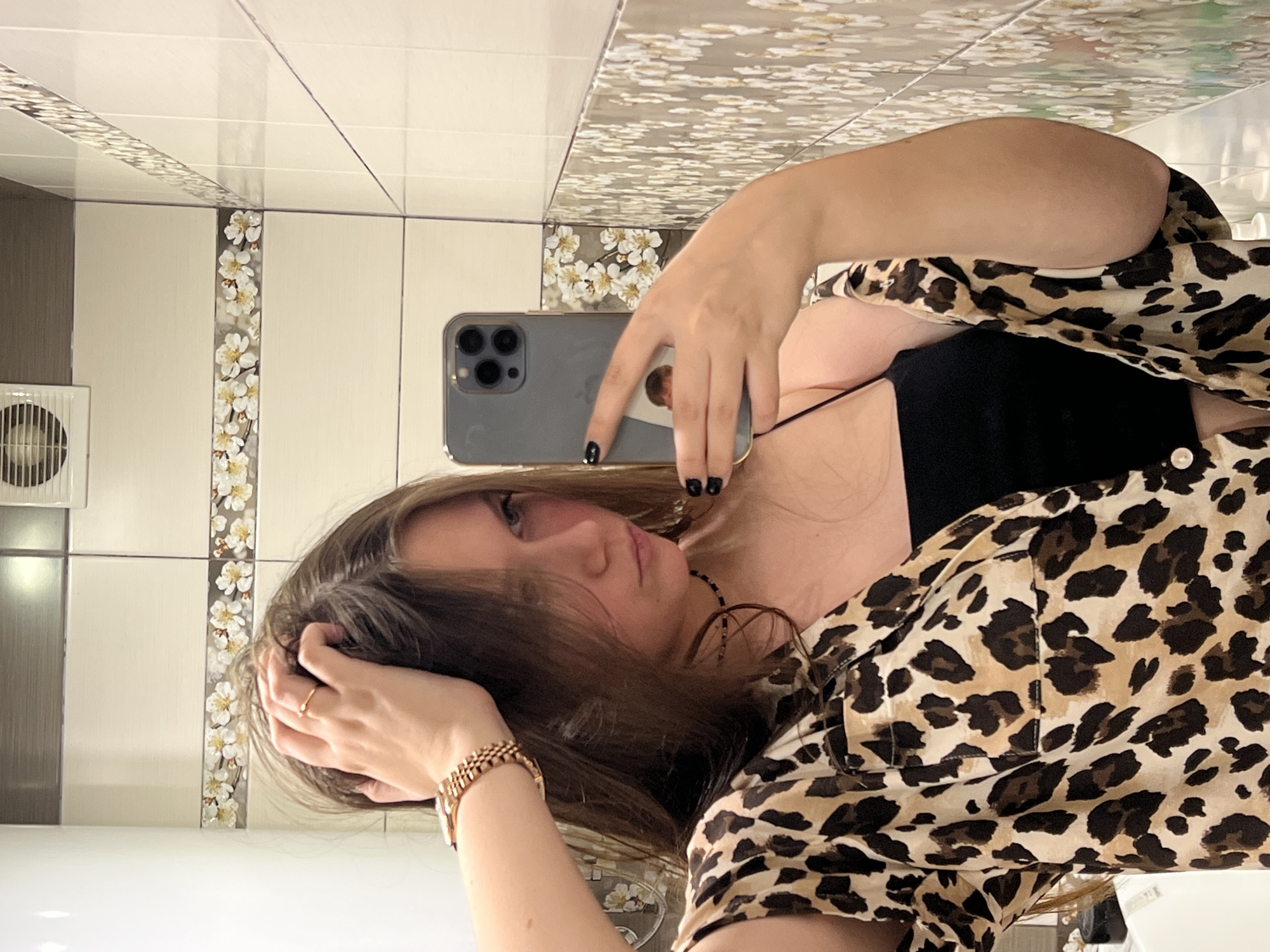 A young woman is taking a selfie in a bathroom. She is wearing a leopard-print shirt over a black top. She is holding a gray smartphone with three camera lenses in her right hand, while her left hand is in her hair. She is wearing a gold-colored bracelet and a ring on her left hand. The bathroom has white tiles with a floral pattern strip. There's a small ventilation fan on the left wall. The location is Chaikovsky, Russia.